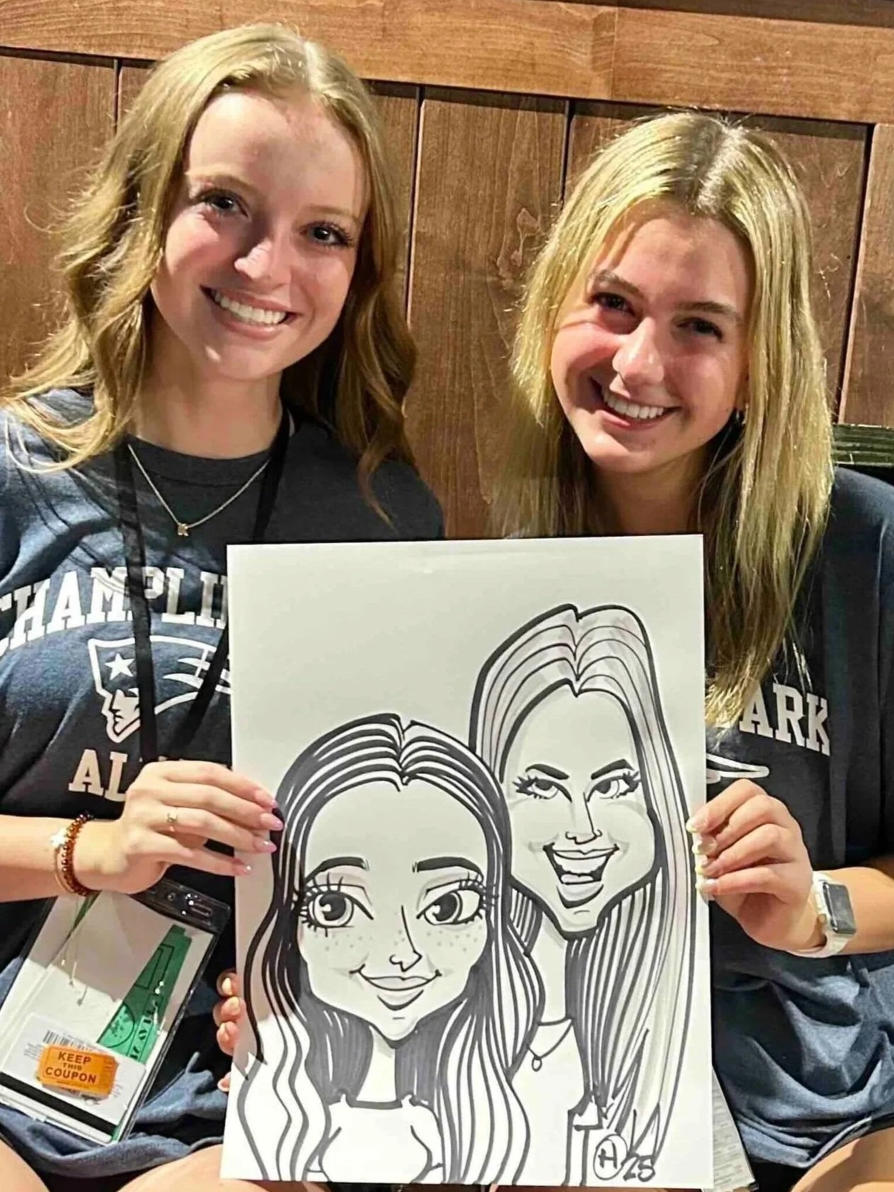 Two smiling women holding a caricature drawing of themselves, with a wooden background at Minneapolis party