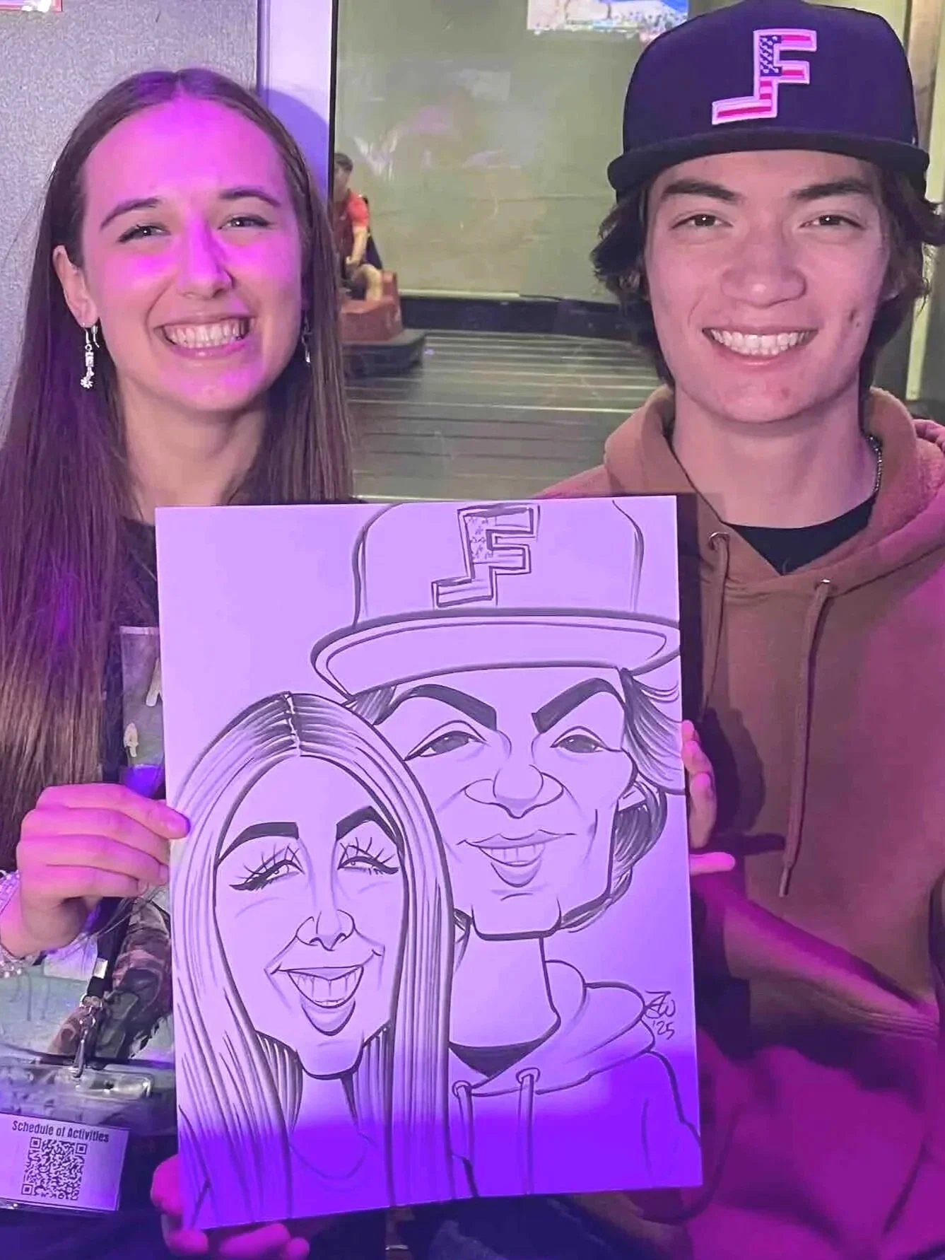 Two people smiling and holding a caricature drawing of themselves. The woman on the left has long hair and earrings, and the man on the right is wearing a cap. The caricature features exaggerated facial features and depicts both individuals smiling.