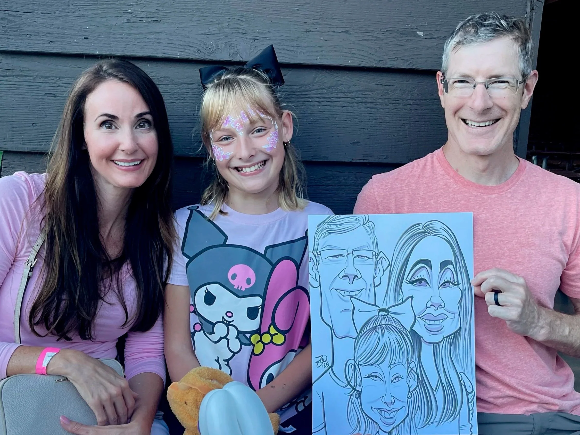 A smiling woman with long brown hair, a young girl with face paint and a black bow in her hair, and a smiling man with glasses holding caricature portraits of themselves. They are standing in front of a dark wooden wall, at Minneapolis event