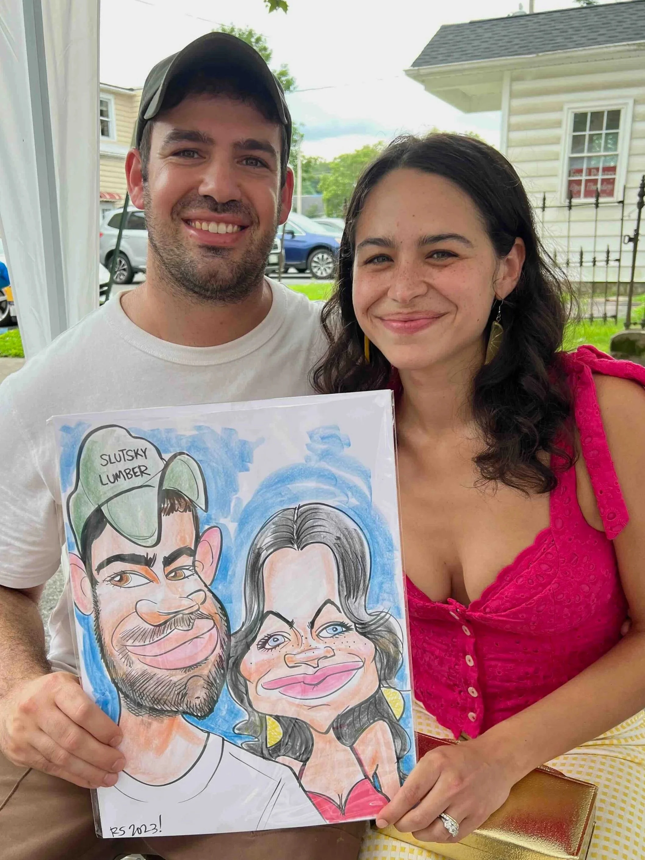 Couple outdoors holding their caricature drawing at a summer event, man in a baseball cap and woman in pink, with a neighborhood backdrop.