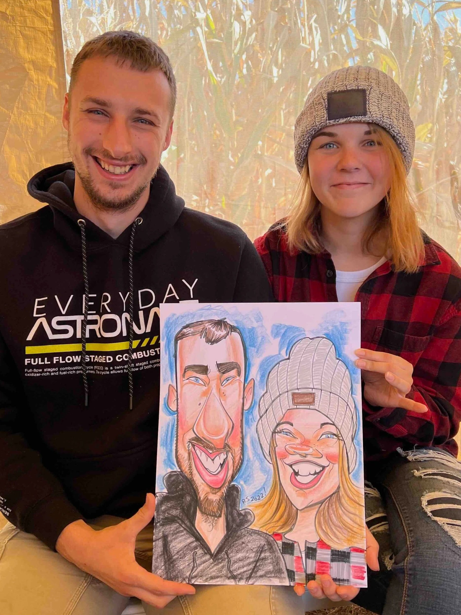 Young couple holding a colorful caricature drawing of themselves at an outdoor event, she in a flannel and beanie, he in a black hoodie.