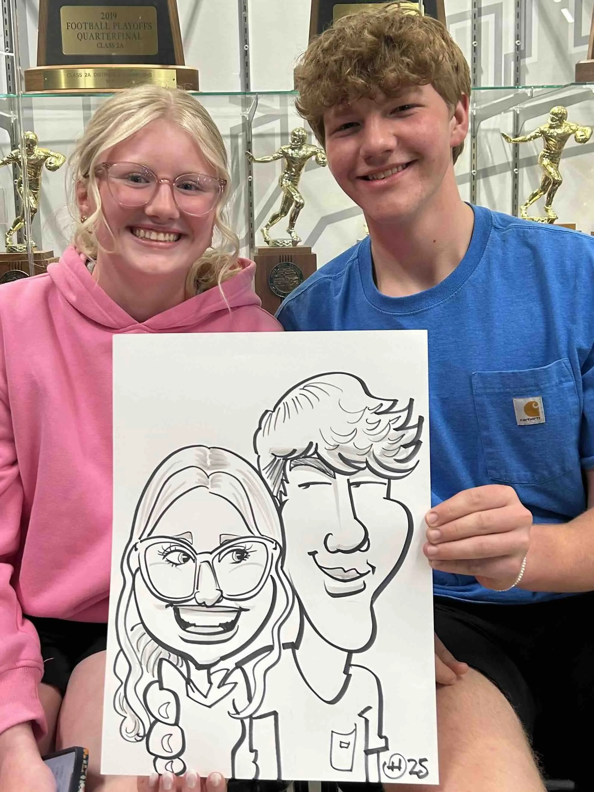 Young woman in a pink hoodie and glasses with a young man in a blue shirt, holding their caricature drawing in front of a sports trophy display at Minnesota high school senior overnight party
