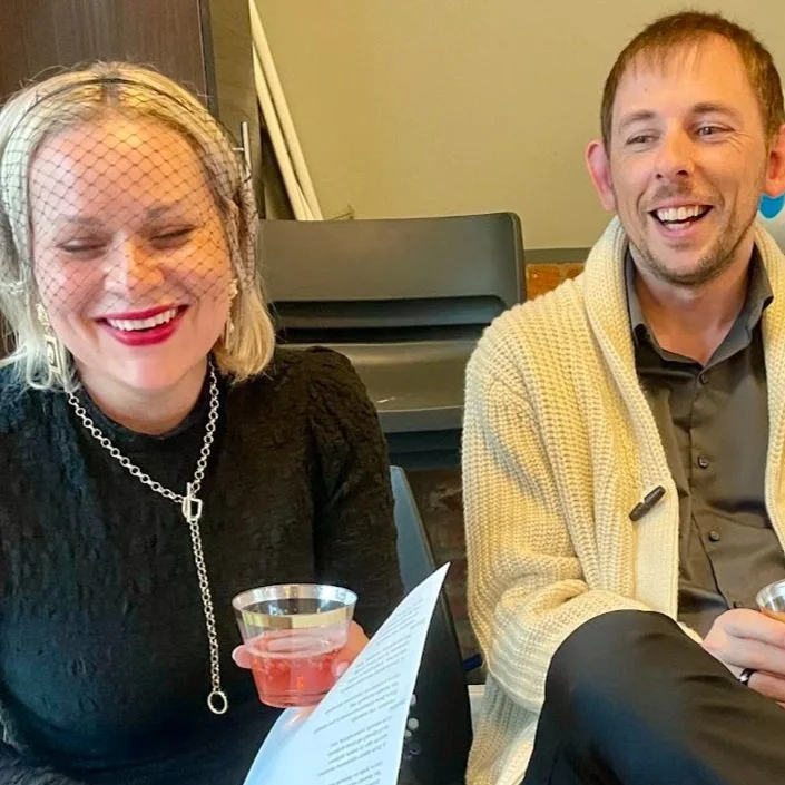 Woman in a black dress and pearl necklace and her companion smiling at a caricature event, holding drinks at a seated venue.