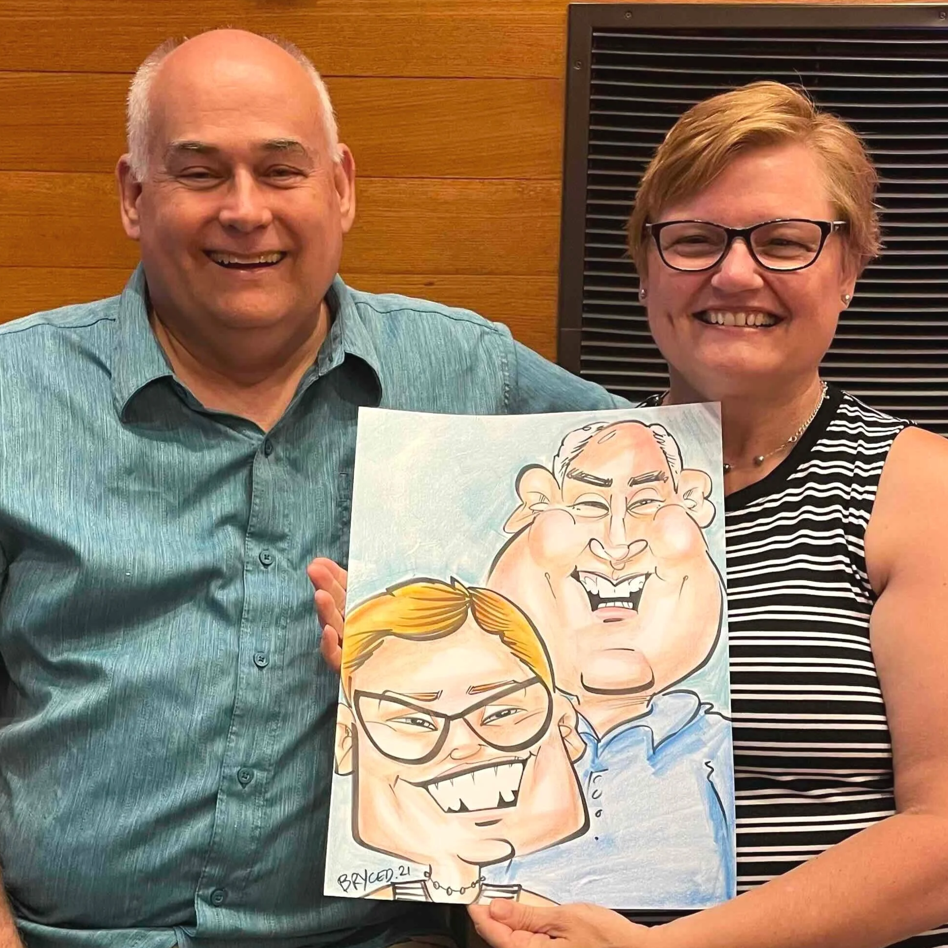 Man and woman smiling and holding their caricature portrait drawn live at a Twin Cities event, with a warm wooden wall backdrop.