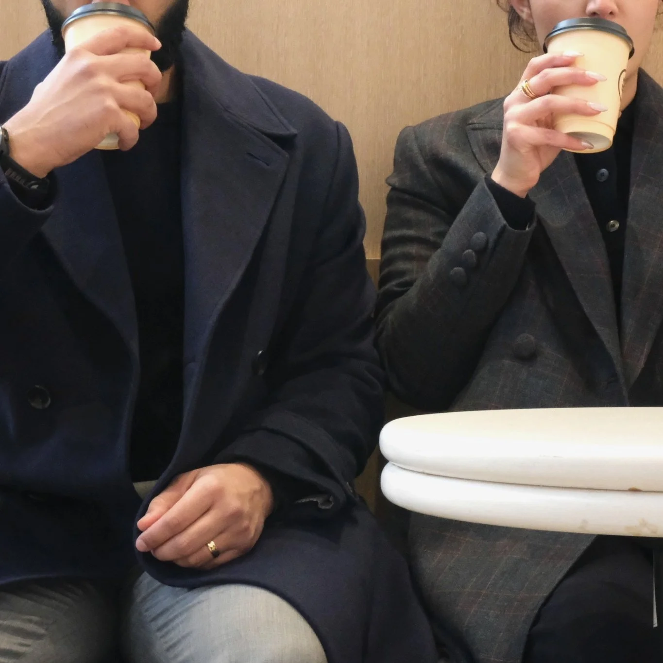 Two people are sitting at a table, each holding a paper cup and drinking from it. They are wearing dark jackets and sitting against a beige wall.