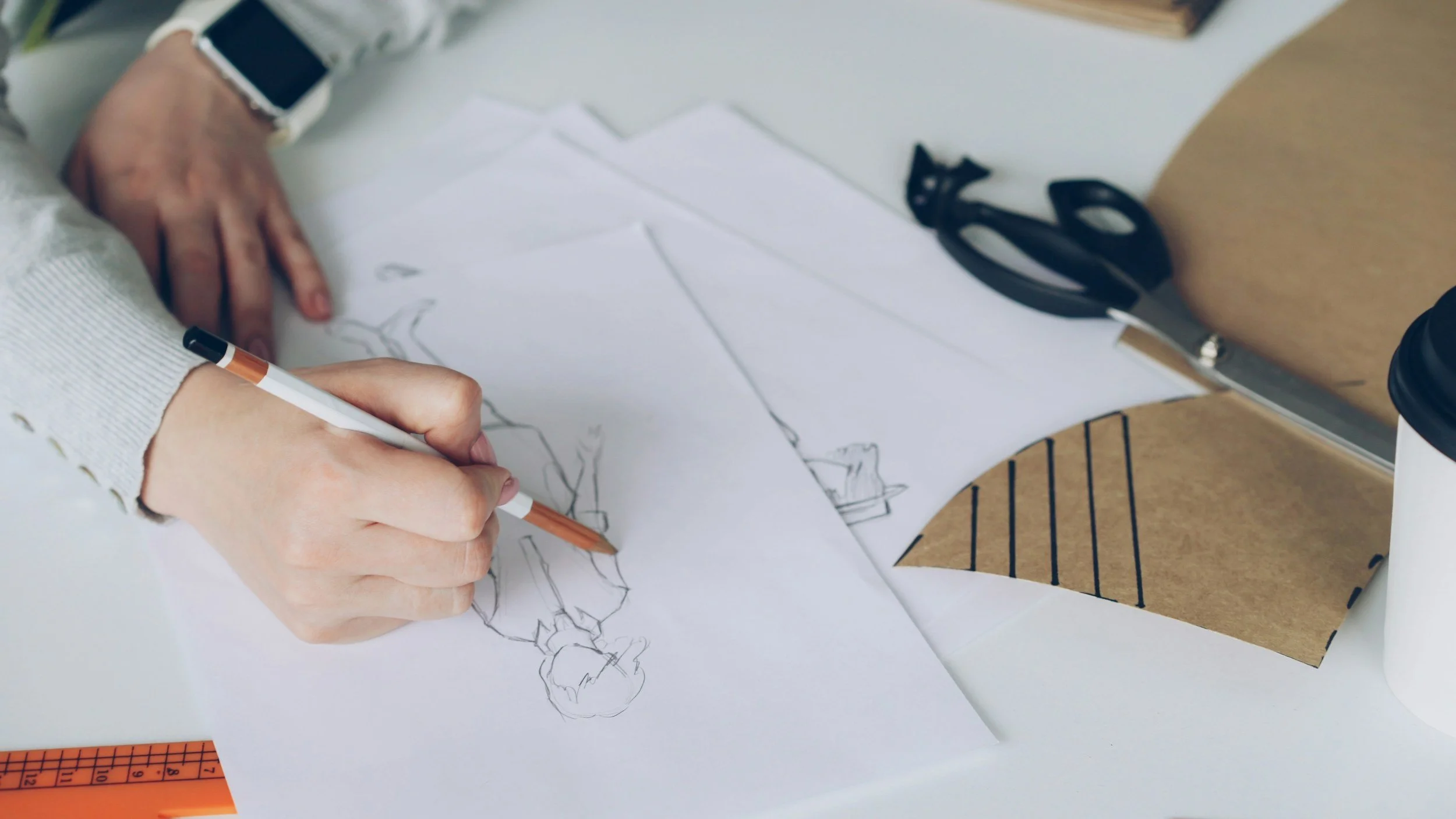 Person sketching a fashion design on paper with tools and fabric scraps nearby.