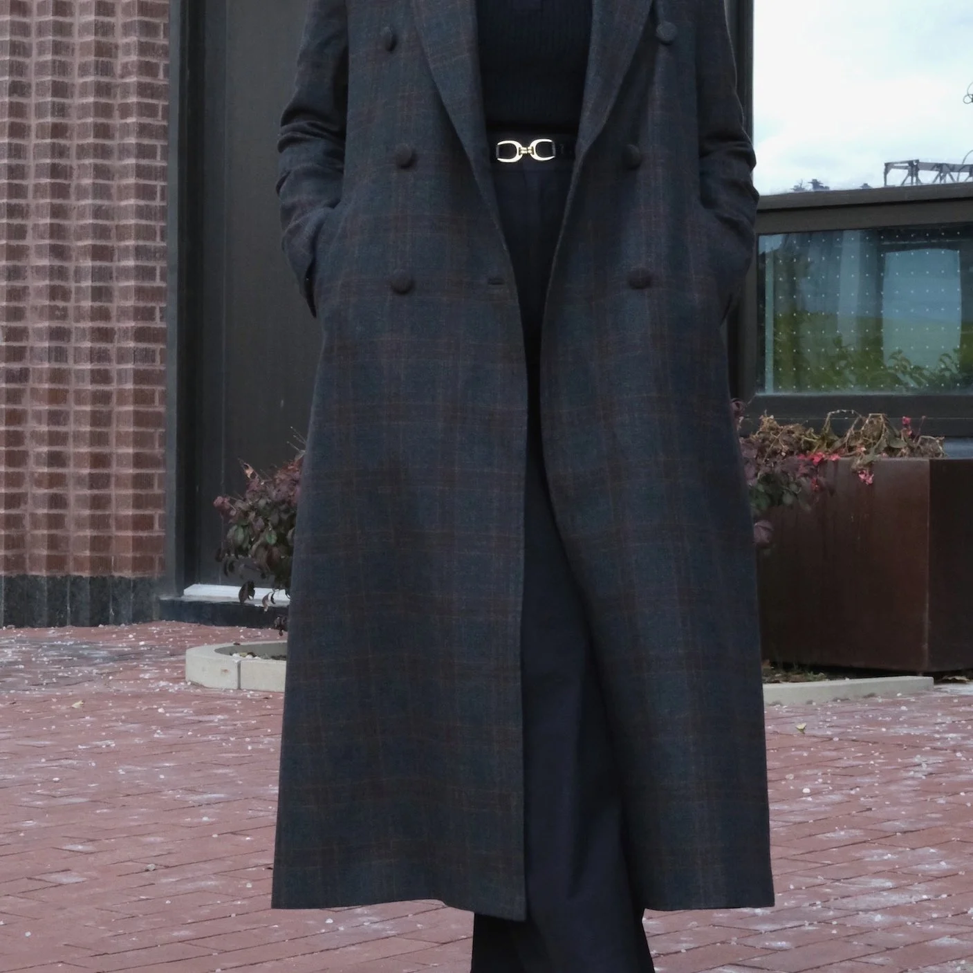 Person wearing a long plaid overcoat with hands in pockets, standing outdoors on a brick pavement.