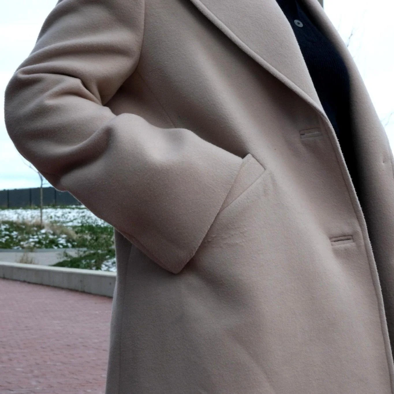 Close-up of a person wearing a beige coat with a pocket, standing outdoors on a cloudy day with snow on the ground in the background.