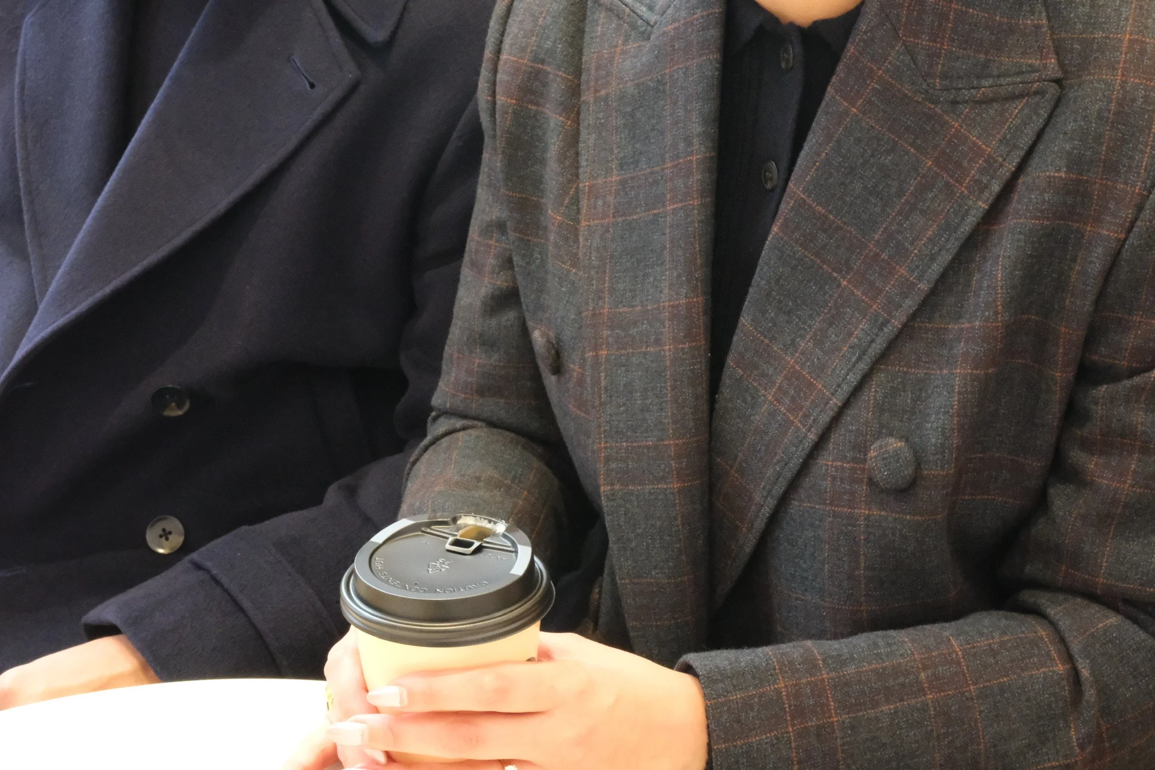 Person in a plaid blazer holding a disposable coffee cup with a black lid, sitting indoors.