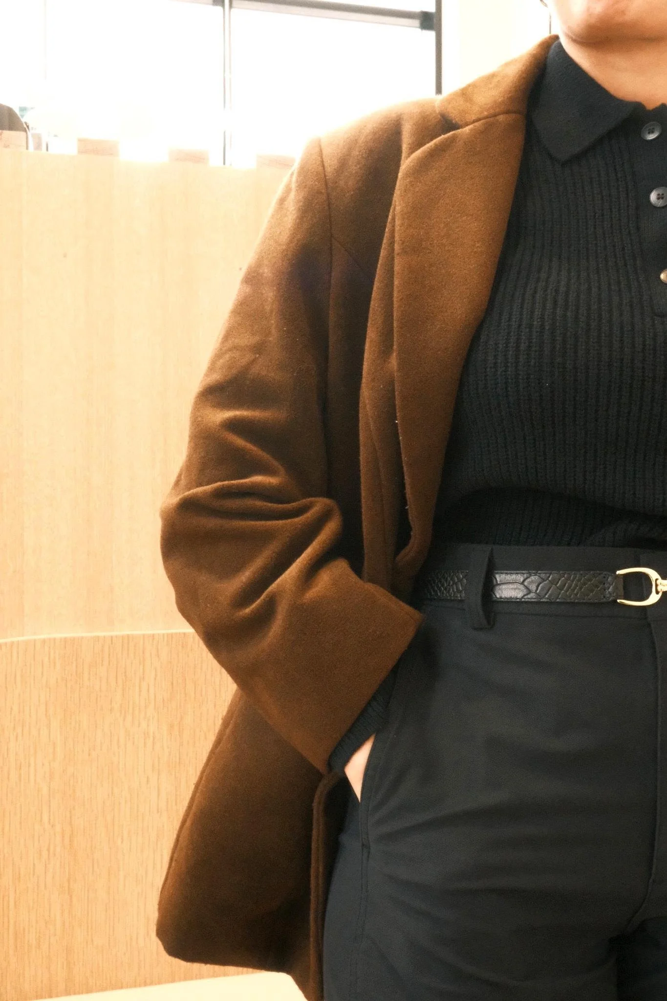 Person wearing a brown coat, black ribbed shirt, black pants, and a black belt with a gold buckle, in front of a wooden panel indoors.