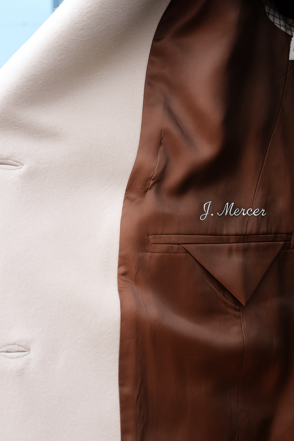 Close-up of a brown satin interior jacket with the embroidered name "J. Mercer" visible, partially covering a cream-colored fabric.