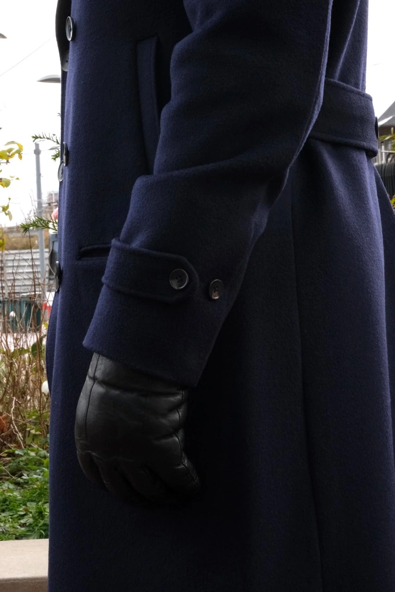 Close-up of a person wearing a navy wool coat, black leather glove, and standing outdoors with some plants and a fence in the background.