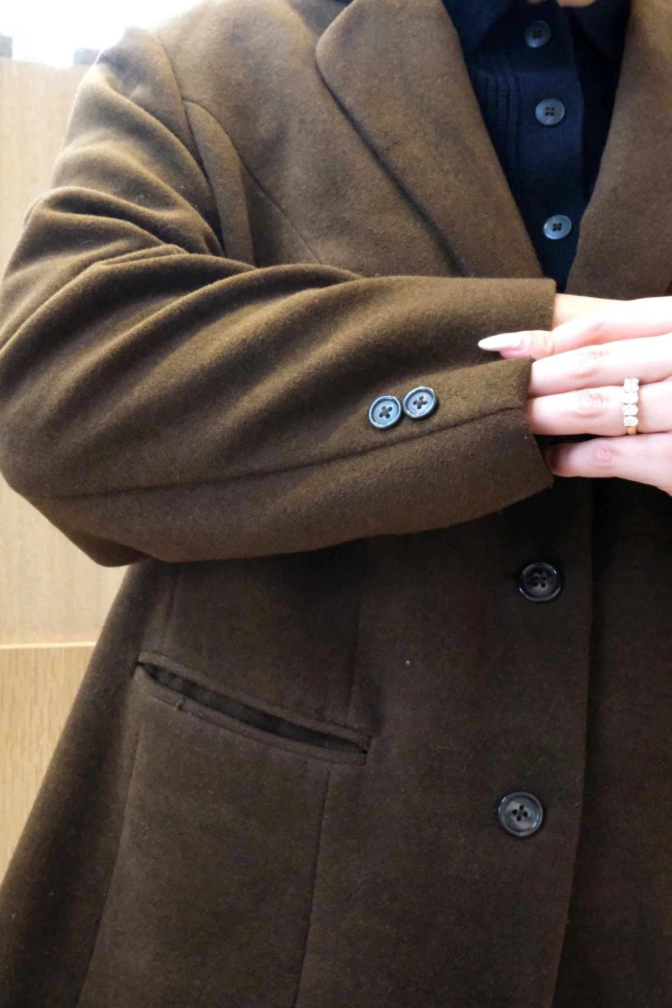 Close-up of a person wearing a brown coat and black shirt, with their hand visible showing a ring on their finger.