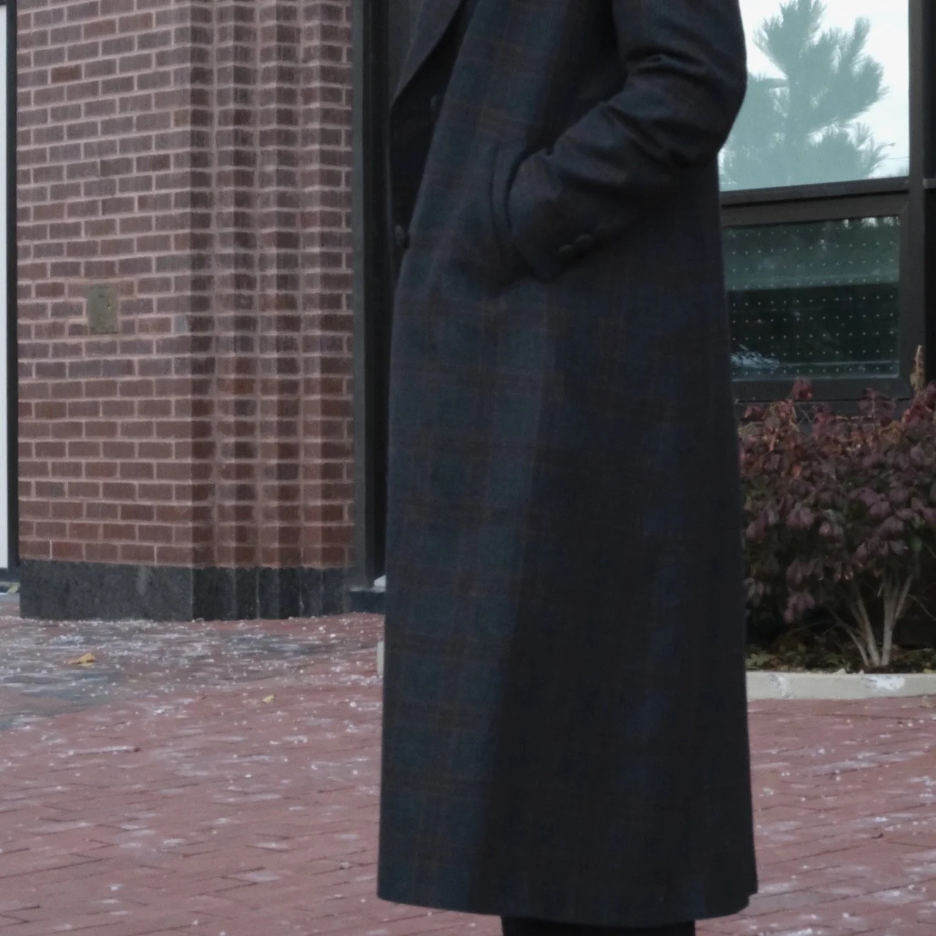 Person wearing a long dark coat standing outside near a brick building with a window and plants.