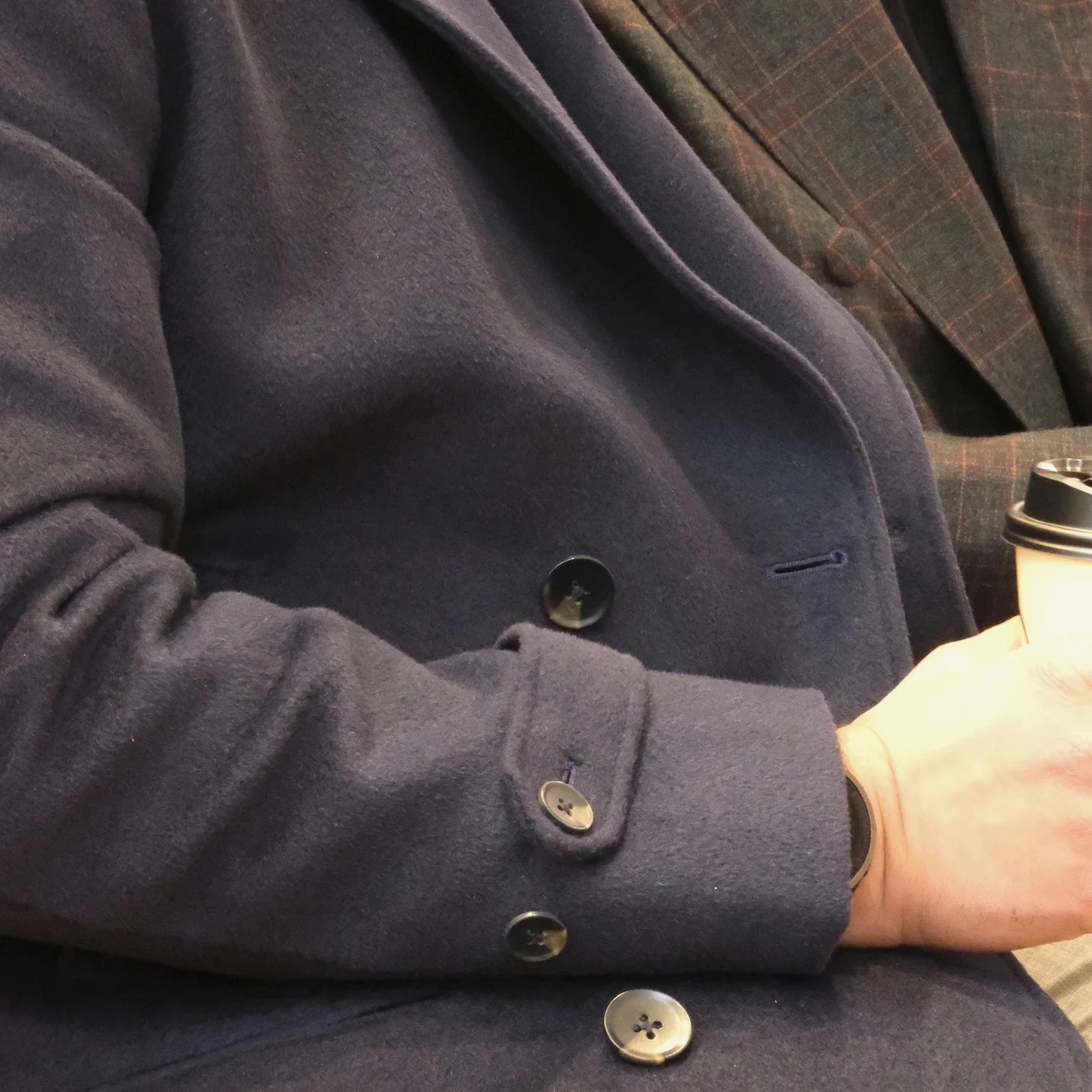 A person wearing a dark coat with buttons, holding a disposable coffee cup, sitting next to another person dressed in a plaid blazer.