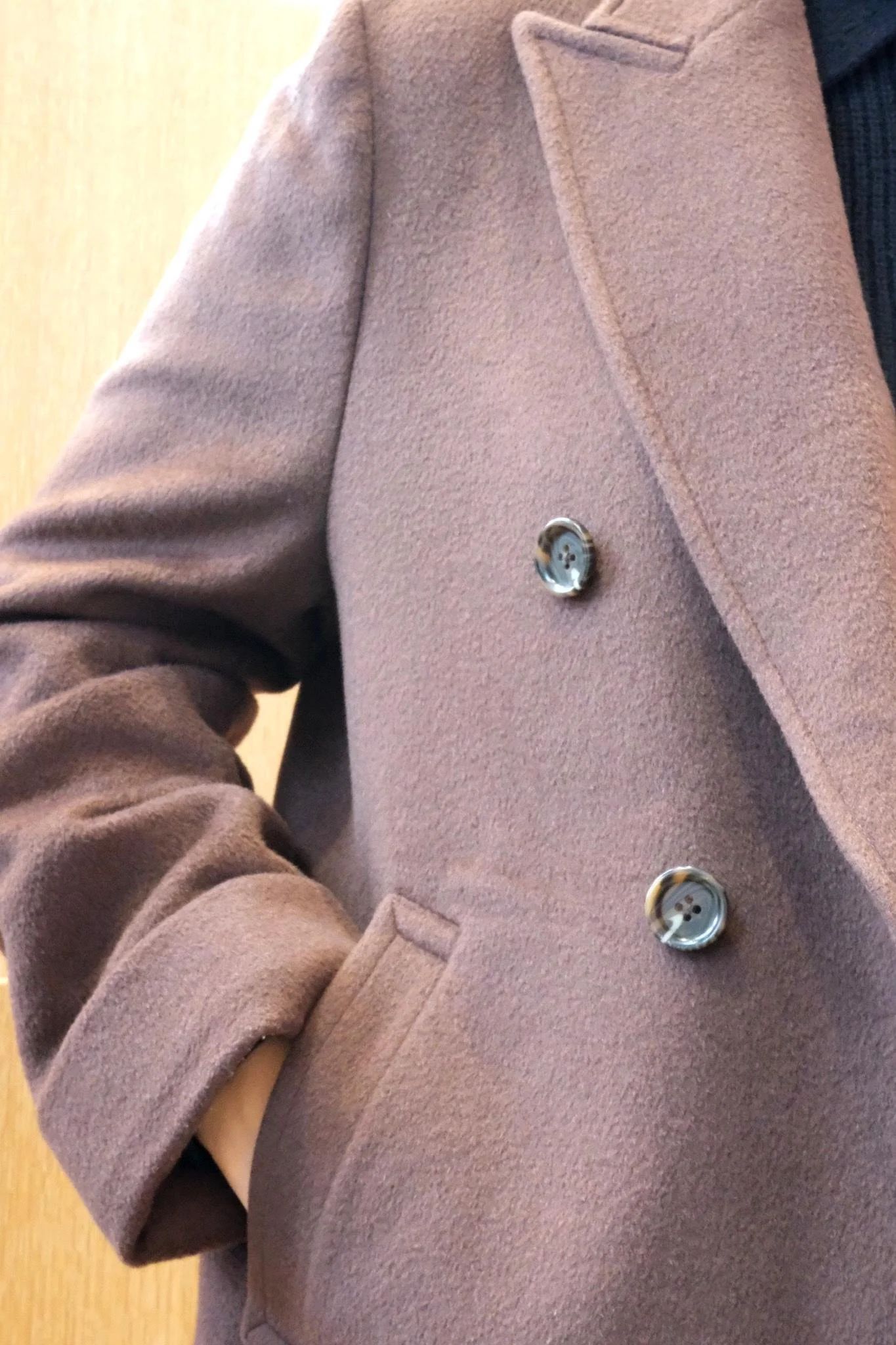 Close-up of a person wearing a light brown coat with the person's hand in the pocket, showing part of a dark shirt underneath.