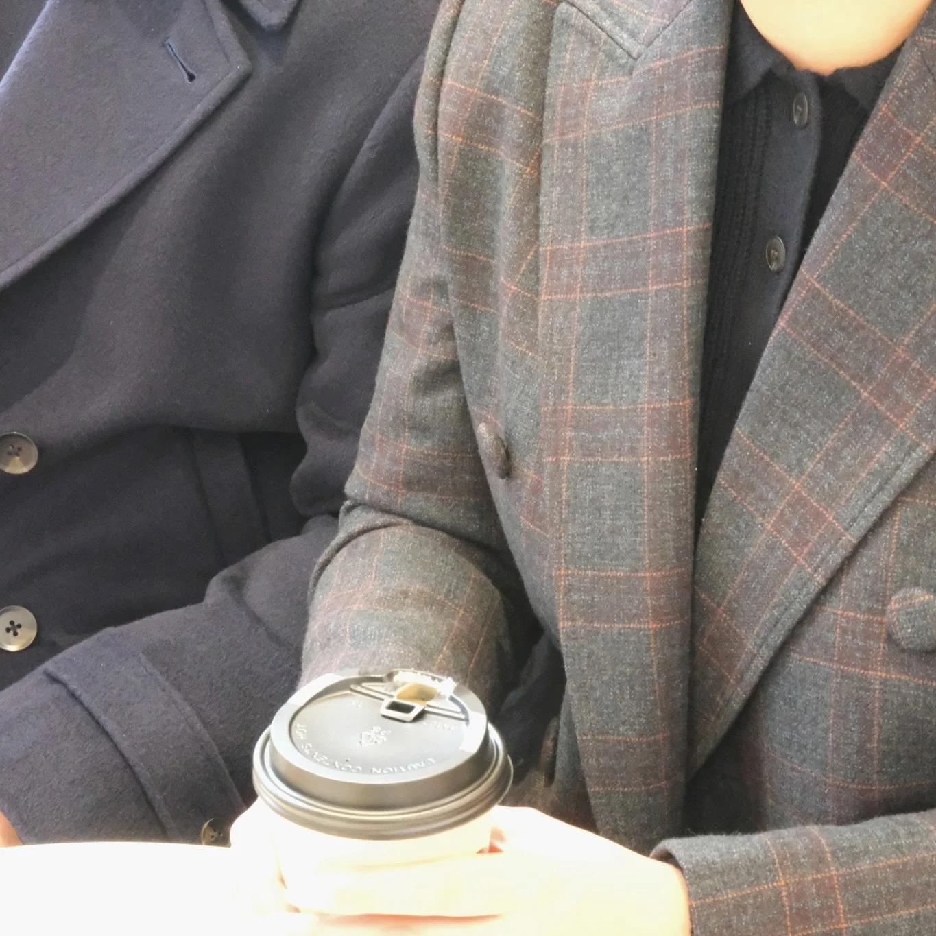 Two people sitting side by side, one wearing a dark coat and the other wearing a gray plaid blazer, holding a to-go coffee cup with a black lid.