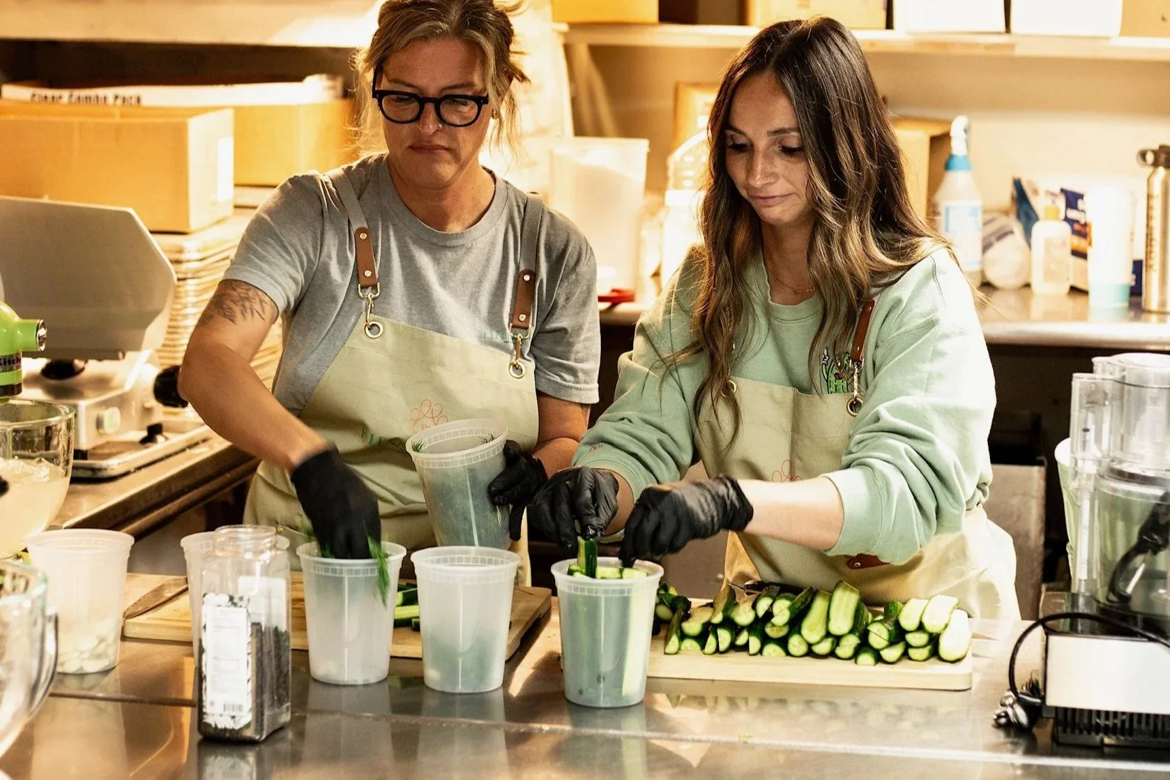 Two women with aprons and black gloves preparing cucumber pieces in a kitchen or craft room with various supplies around them.