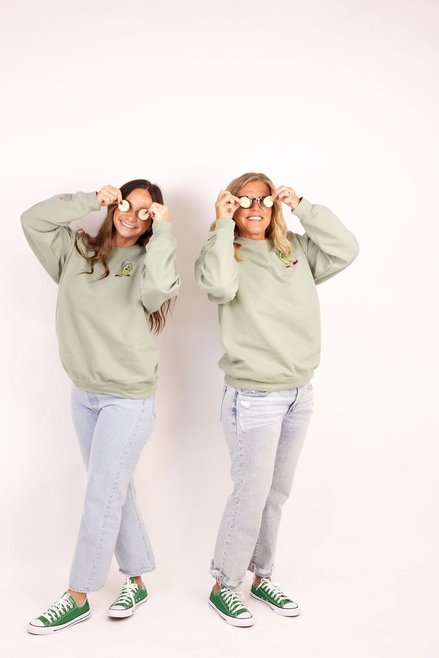 Two women standing against a plain white background, wearing matching gray sweatshirts and light jeans, holding slices of cucumber over their eyes, smiling, with one woman wearing glasses and both wearing green sneakers.