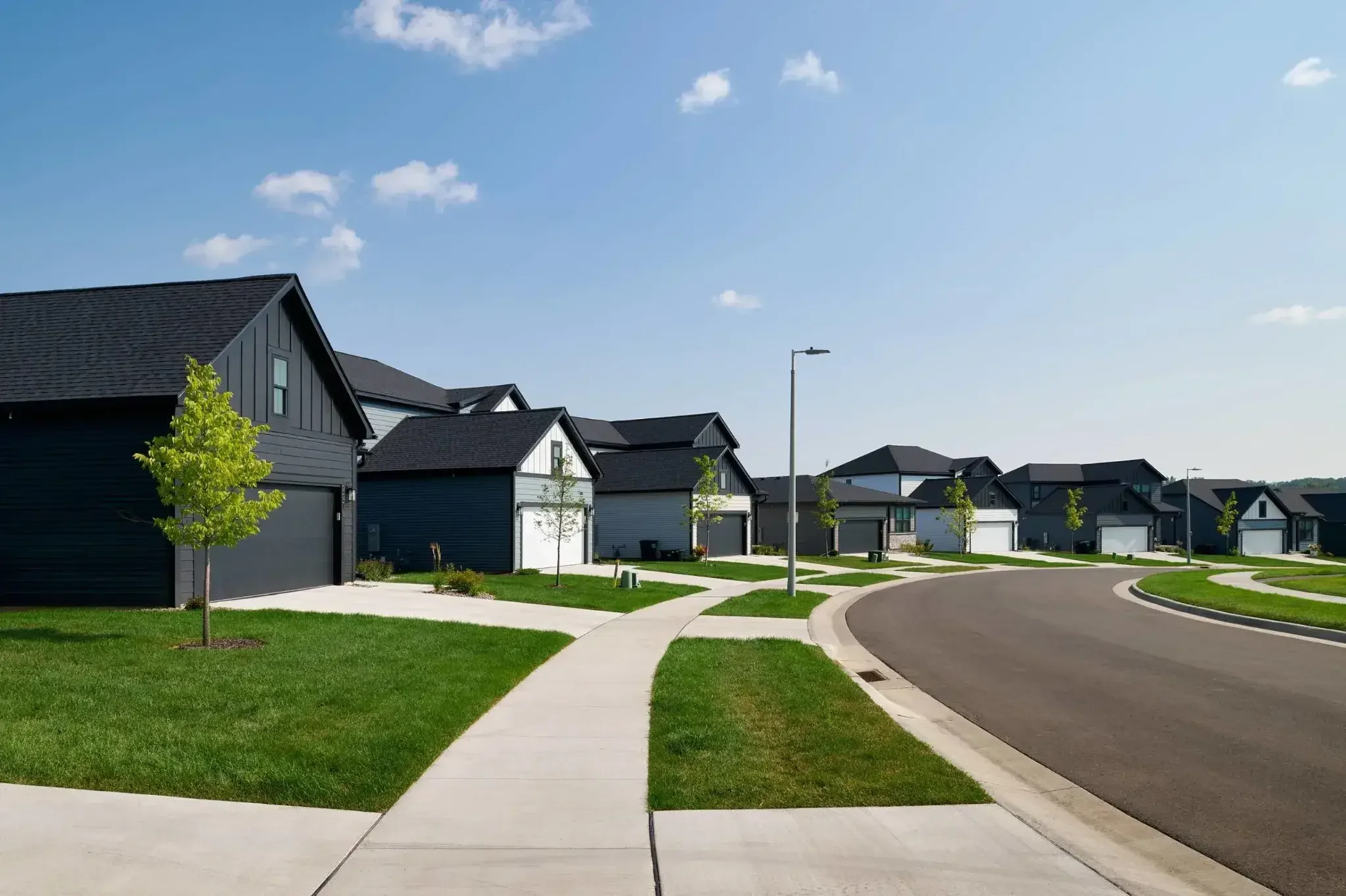 A suburban neighborhood with curved street and sidewalk, modern dark-colored houses with garages, neatly maintained green lawns, small trees, and clear blue sky with a few clouds.