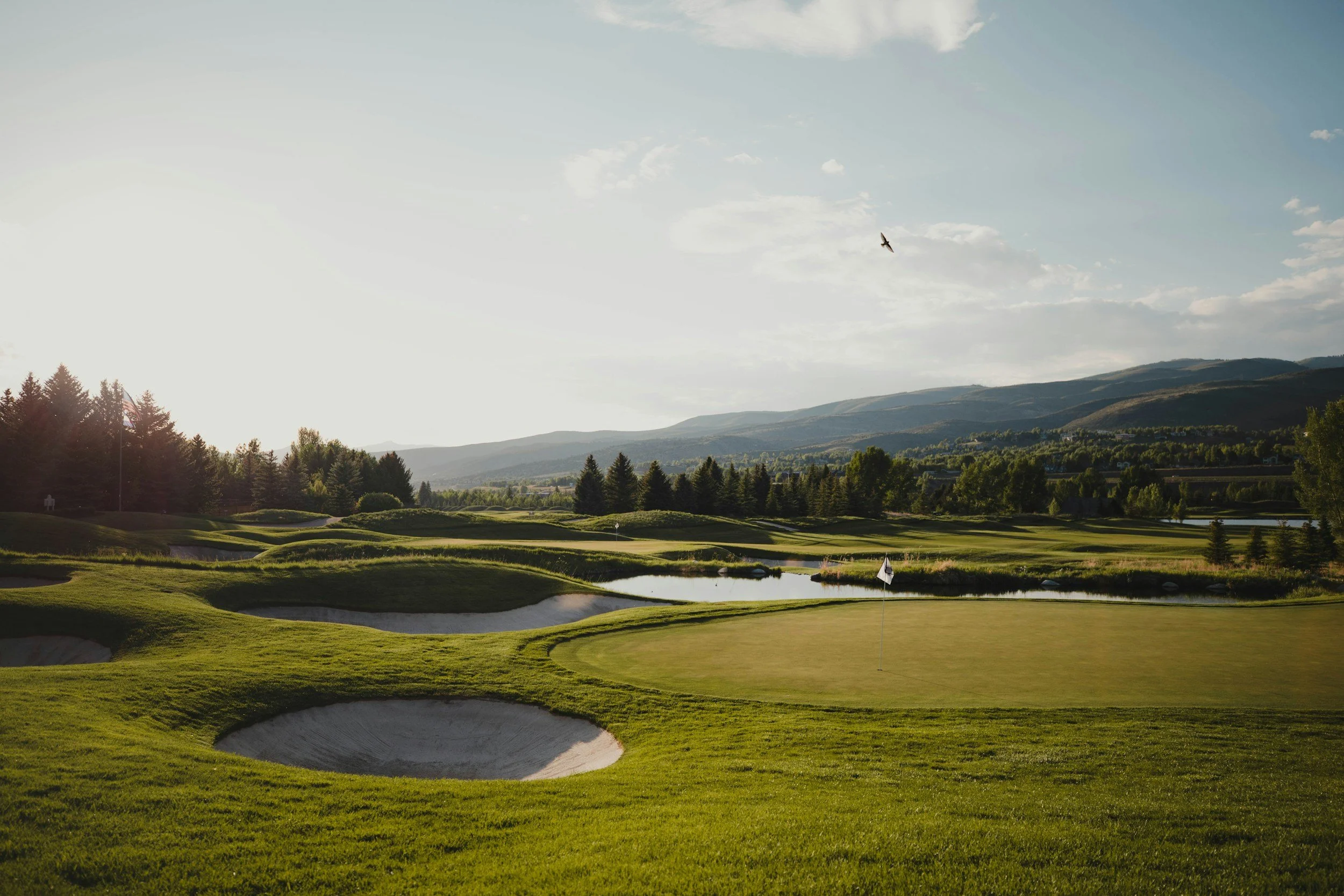 A scenic view of a golf course in the late afternoon or early evening with well-maintained green grass, sand bunkers, and a small water hazard, surrounded by trees and mountains under a partly cloudy sky.