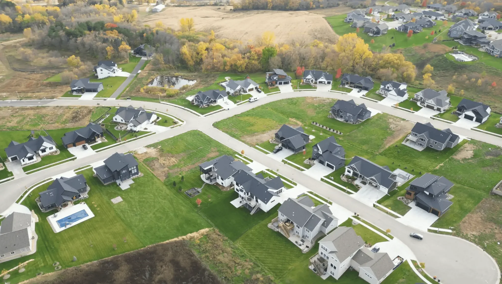 Aerial view of a suburban neighborhood with new houses, green lawns, and a curved street layout during autumn, with some trees displaying fall foliage.