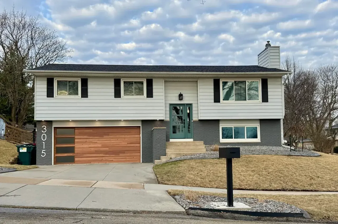 A two-story house with a two-car garage, white siding, black shutters, gray brick accents, and a green front door. The lawn is brown, and there is a black mailbox in front.