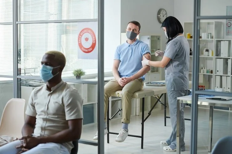 A man in a blue shirt sitting on a medical examination table receiving a vaccination from a nurse in scrubs, with a woman sitting nearby and a man with a face mask sitting in the foreground in a medical clinic.
