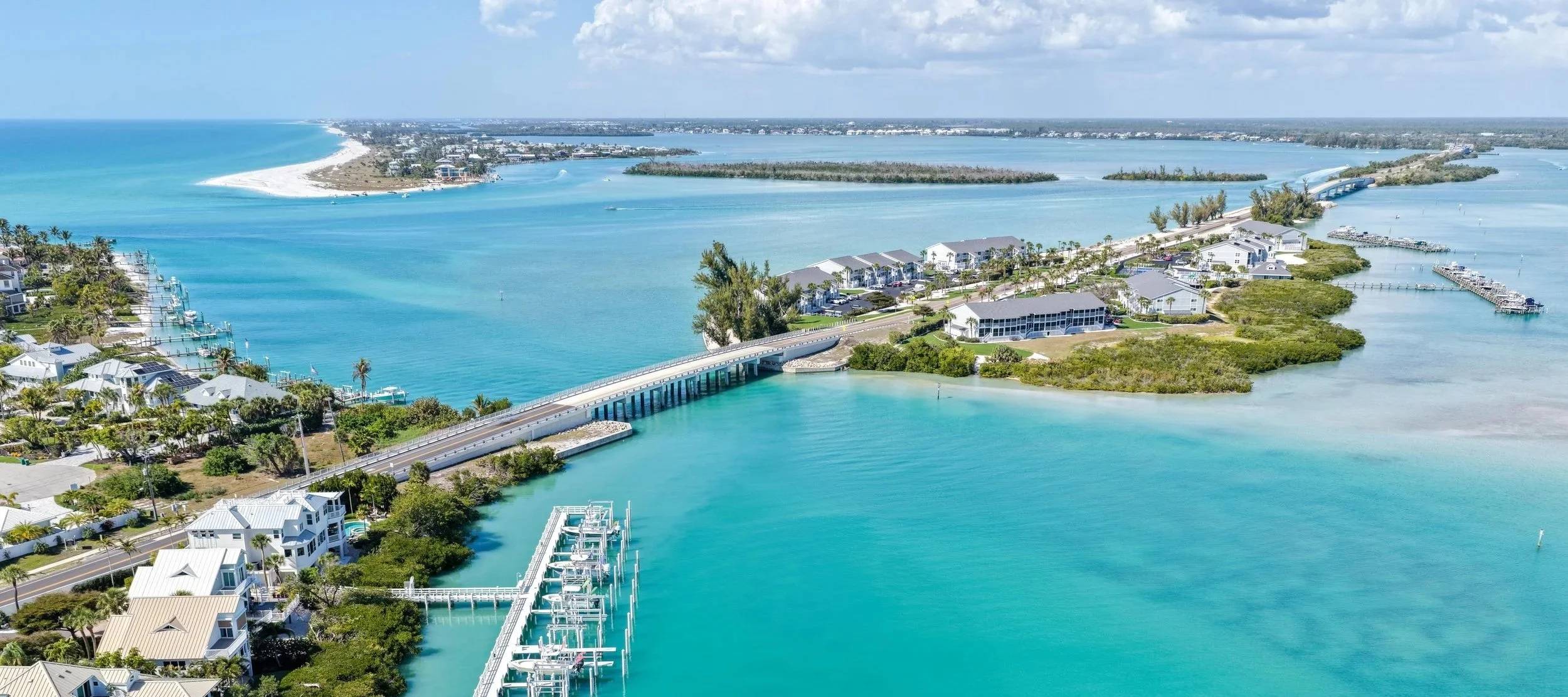 Aerial view of a coastal area with turquoise waters, a bridge, residential buildings, and a marina with boats, set against a backdrop of a beach and distant land.