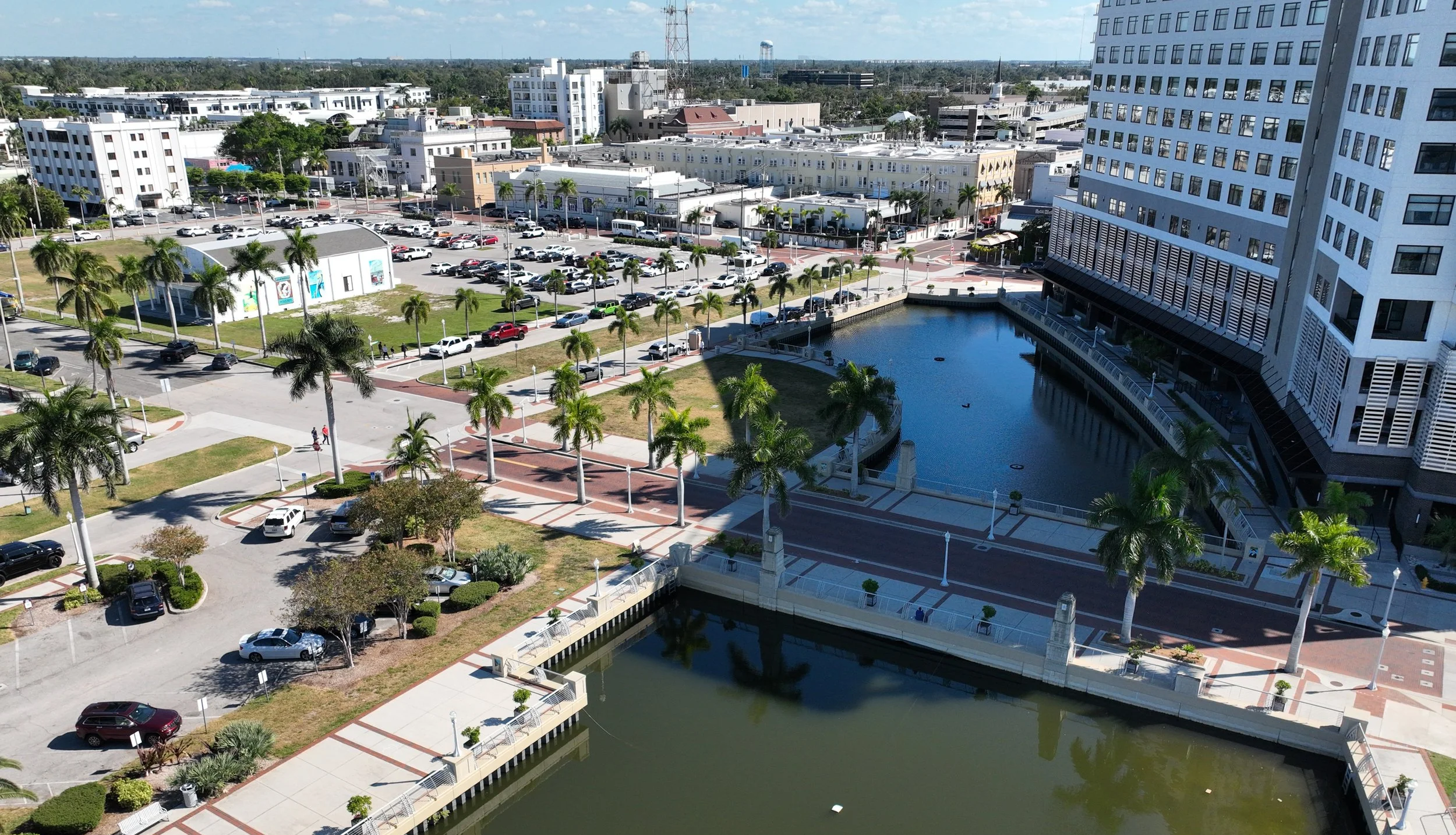 Aerial view of a cityscape featuring a large modern high-rise building, a pond, landscaped areas with palm trees, parking lots filled with cars, and surrounding buildings in the background.