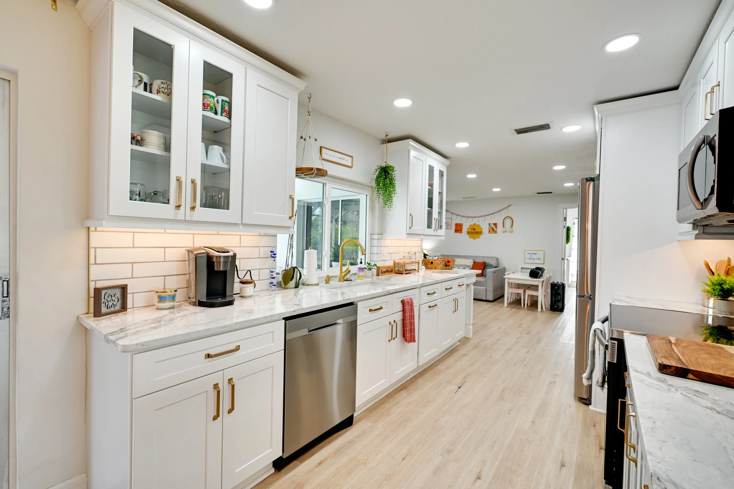 A modern kitchen with white cabinets, marble countertops, gold hardware, stainless steel appliances, and decorative touches. In the background, there is a small living area with a gray sofa and colorful wall art.