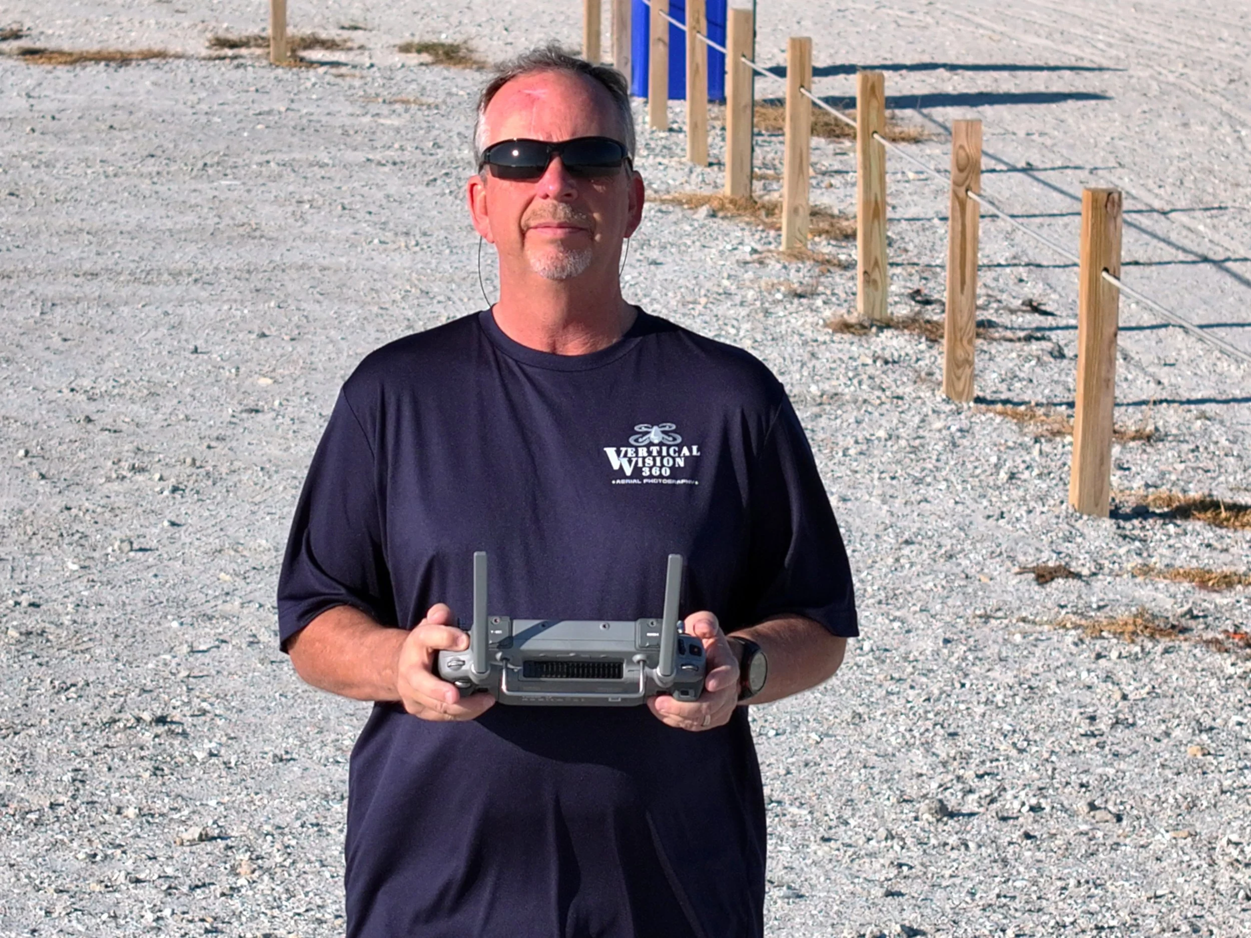 A man wearing sunglasses and a black t-shirt holding a drone controller outdoors on a gravel surface.