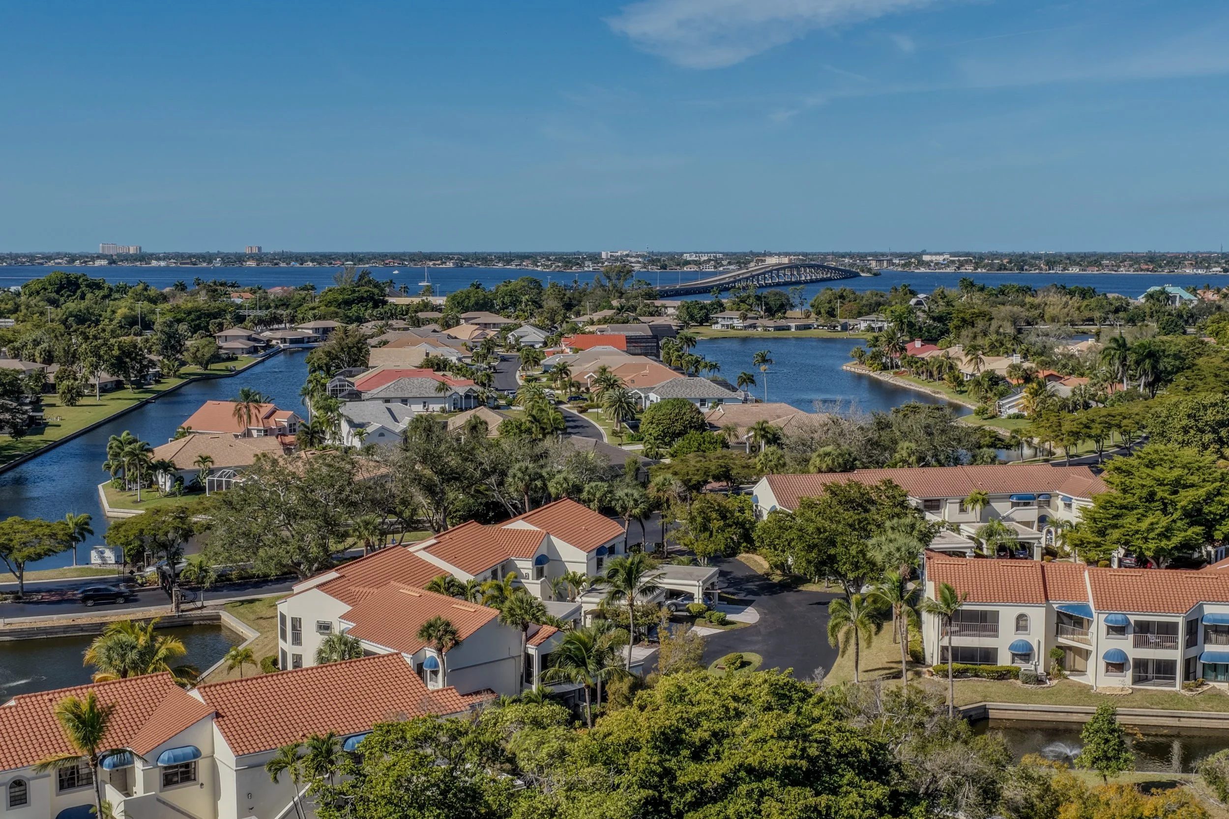 Aerial view of residential neighborhood with houses, waterways, and a bridge in the background under a blue sky.