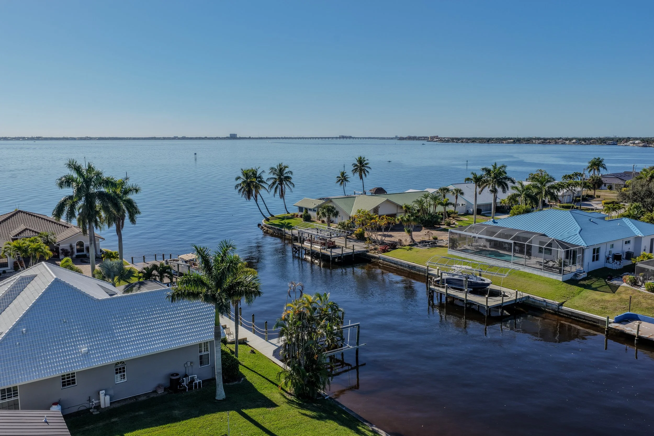 Aerial view of a waterfront neighborhood with houses, docks, and palm trees along a calm river under a clear blue sky.