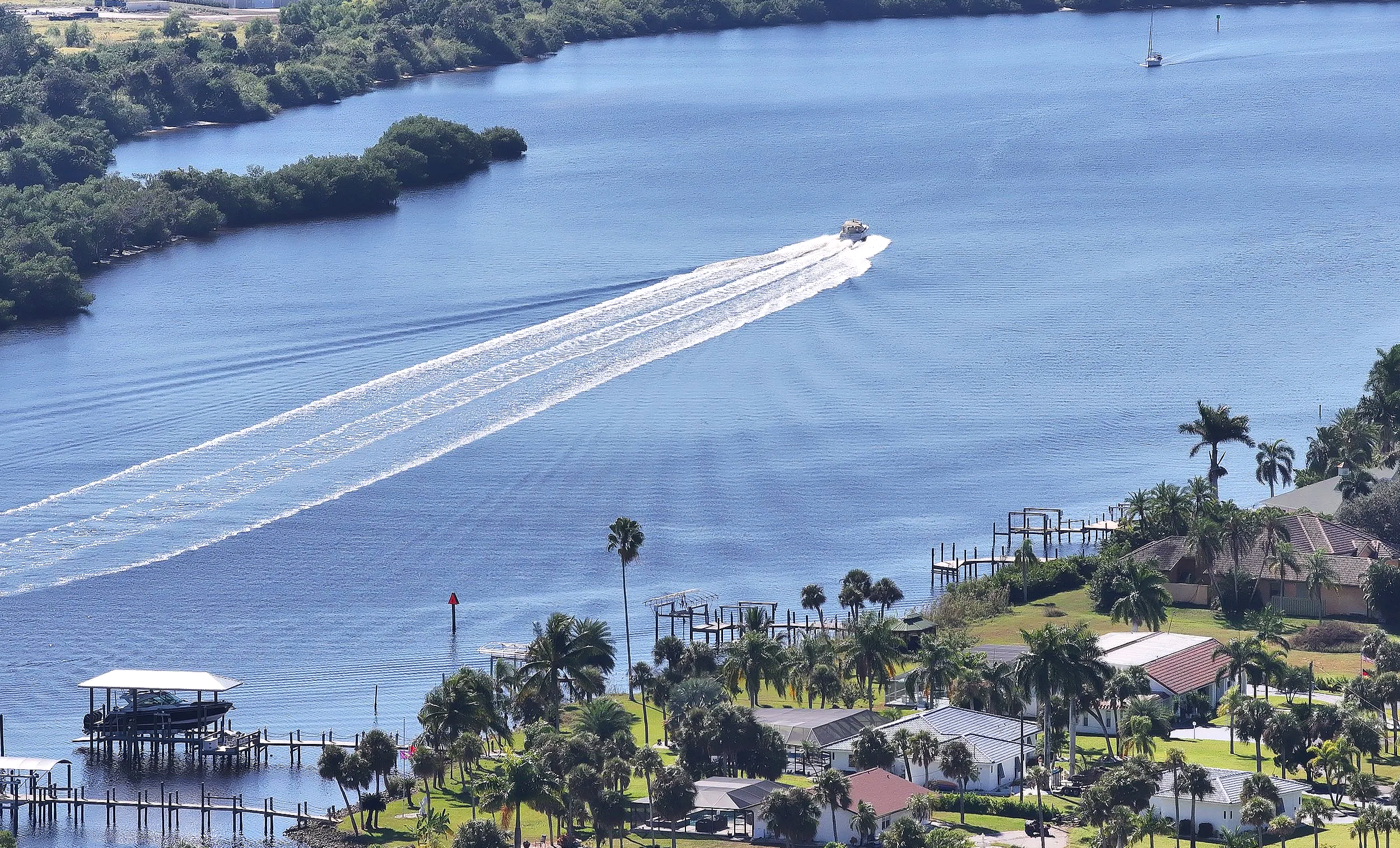 A boat moving on a river, leaving a wake behind it, with houses and trees along the riverbank.