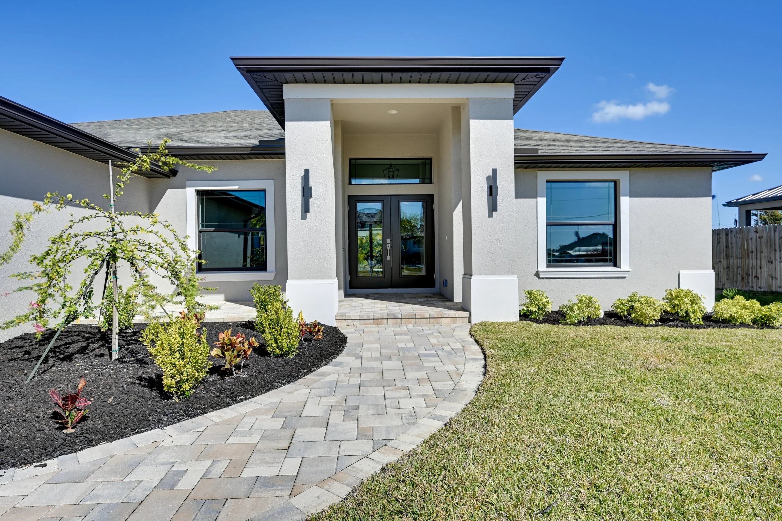 Front view of a modern house with a paved walkway leading to the porch, flanked by well-maintained lawn and landscaped bushes, under a blue sky.