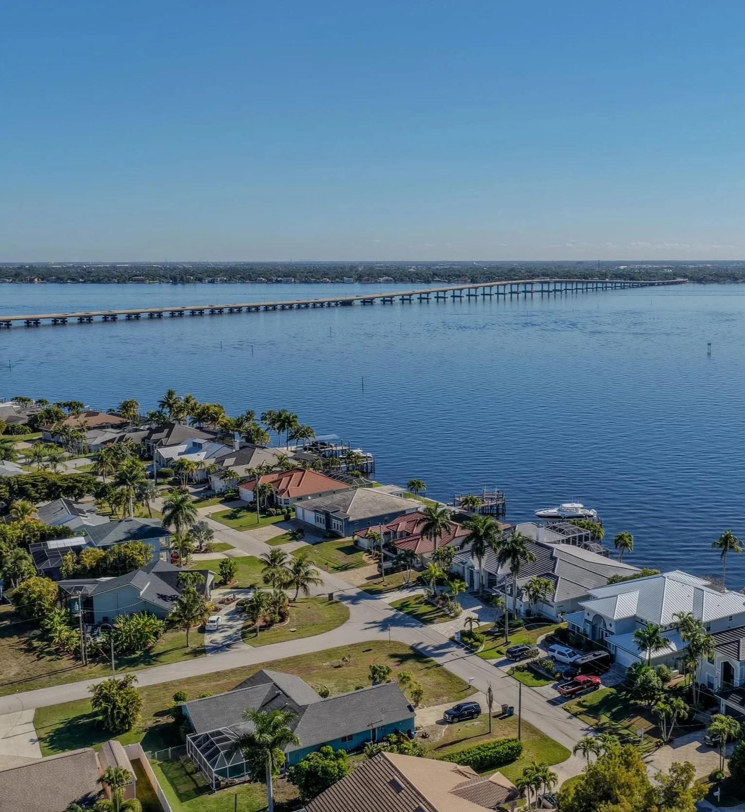 Aerial view of a neighborhood with houses and palm trees along a water body with a long bridge extending across the water under a clear blue sky.