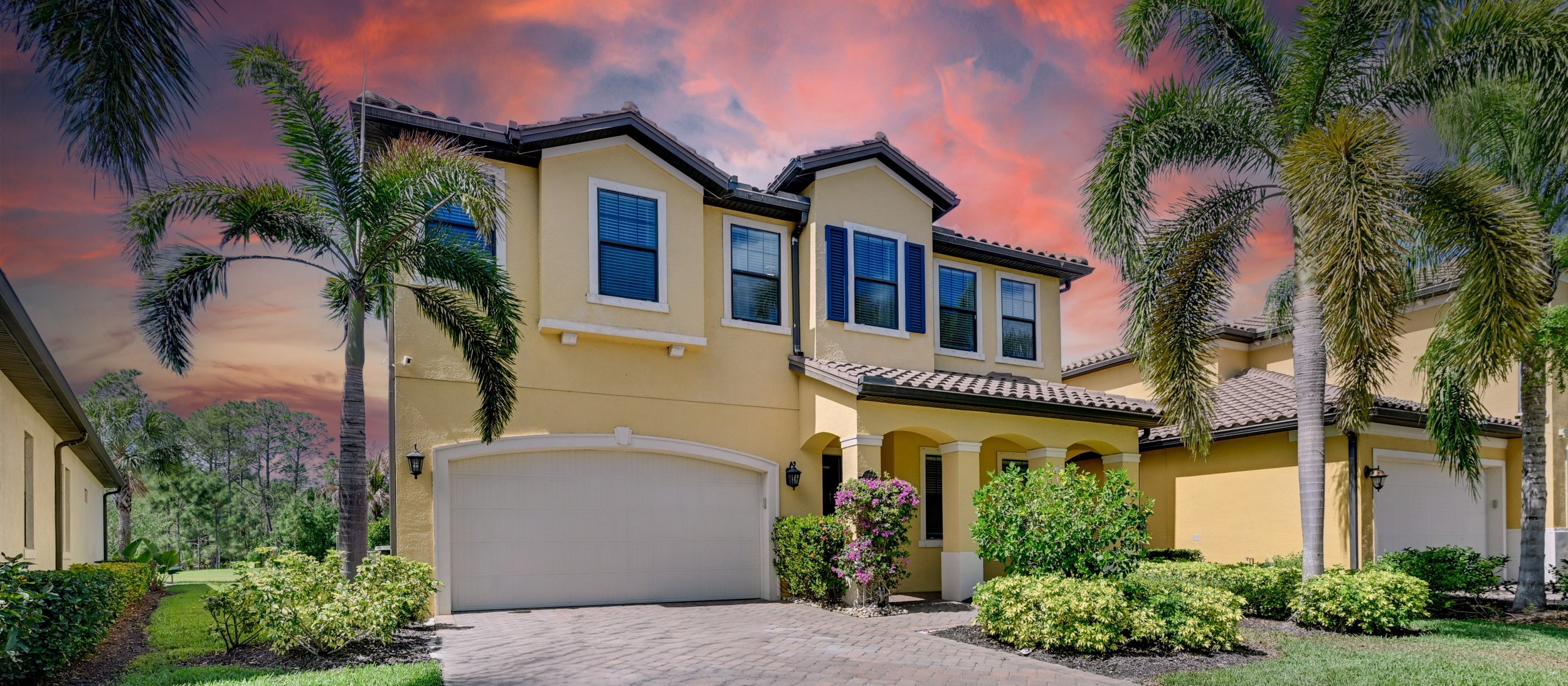 A yellow two-story house with a tiled roof, blue window shutters, and a garage, surrounded by palm trees and well-maintained garden at sunset.