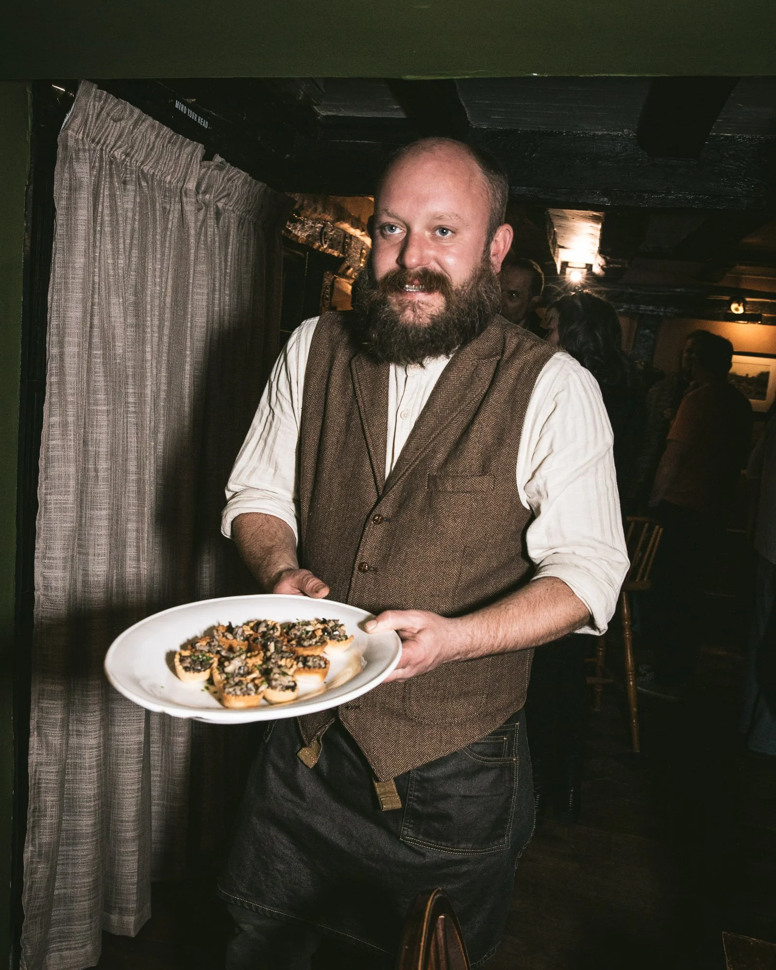 A man with a beard wearing a brown vest and white shirt holding a plate of appetizers in a cozy restaurant or pub setting.