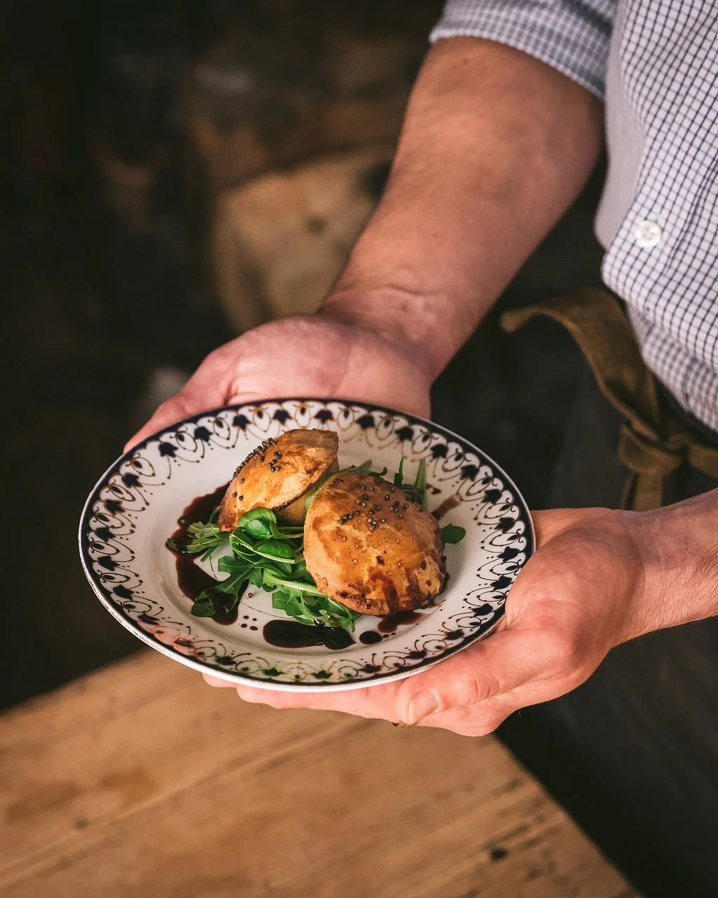 A person holding a decorative plate with two grilled burger patties on a bed of greens, drizzled with sauce.
