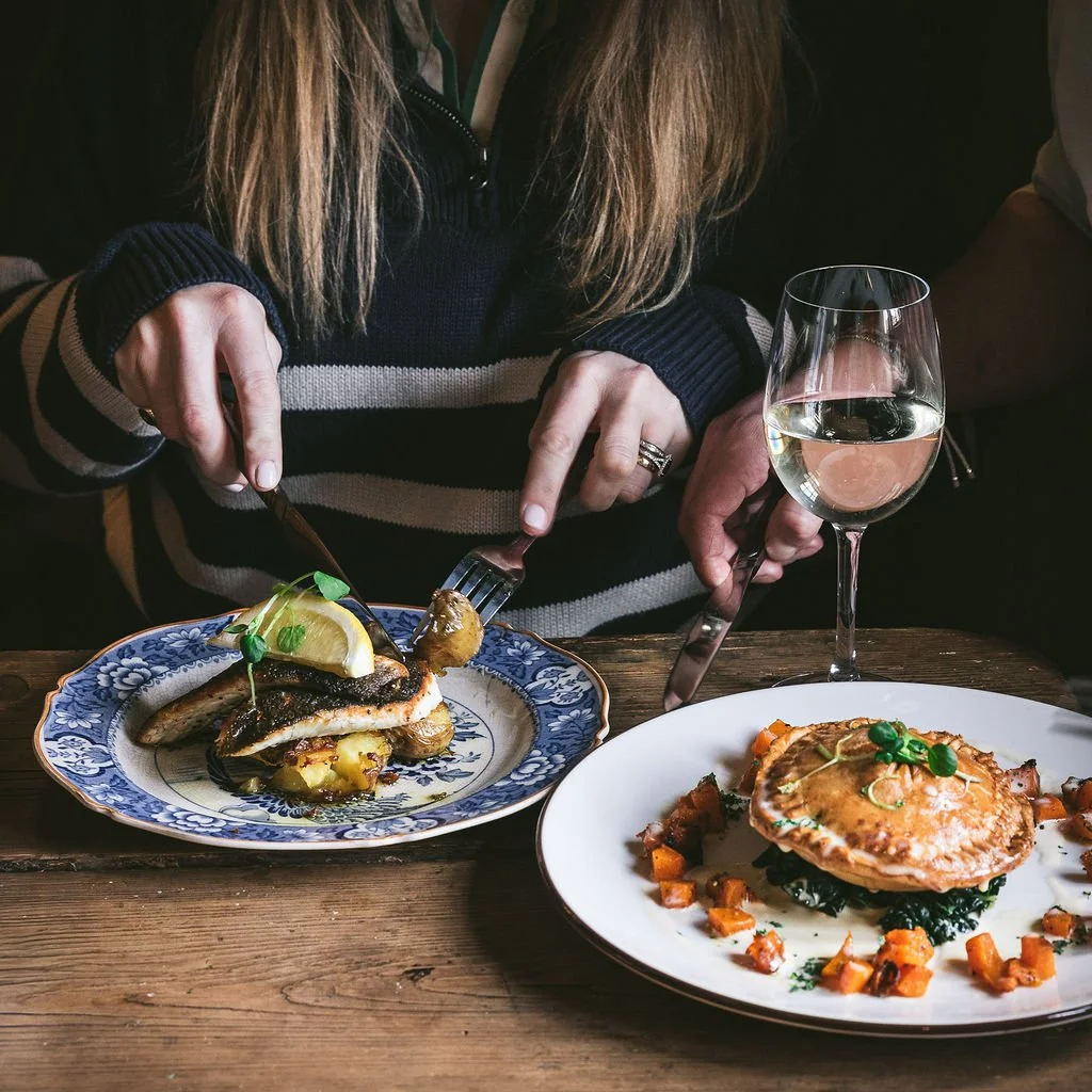 Person serving a grilled fish dish with lemon and herbs at a restaurant table, with a glass of white wine and a plate of baked puff pastry with vegetables.
