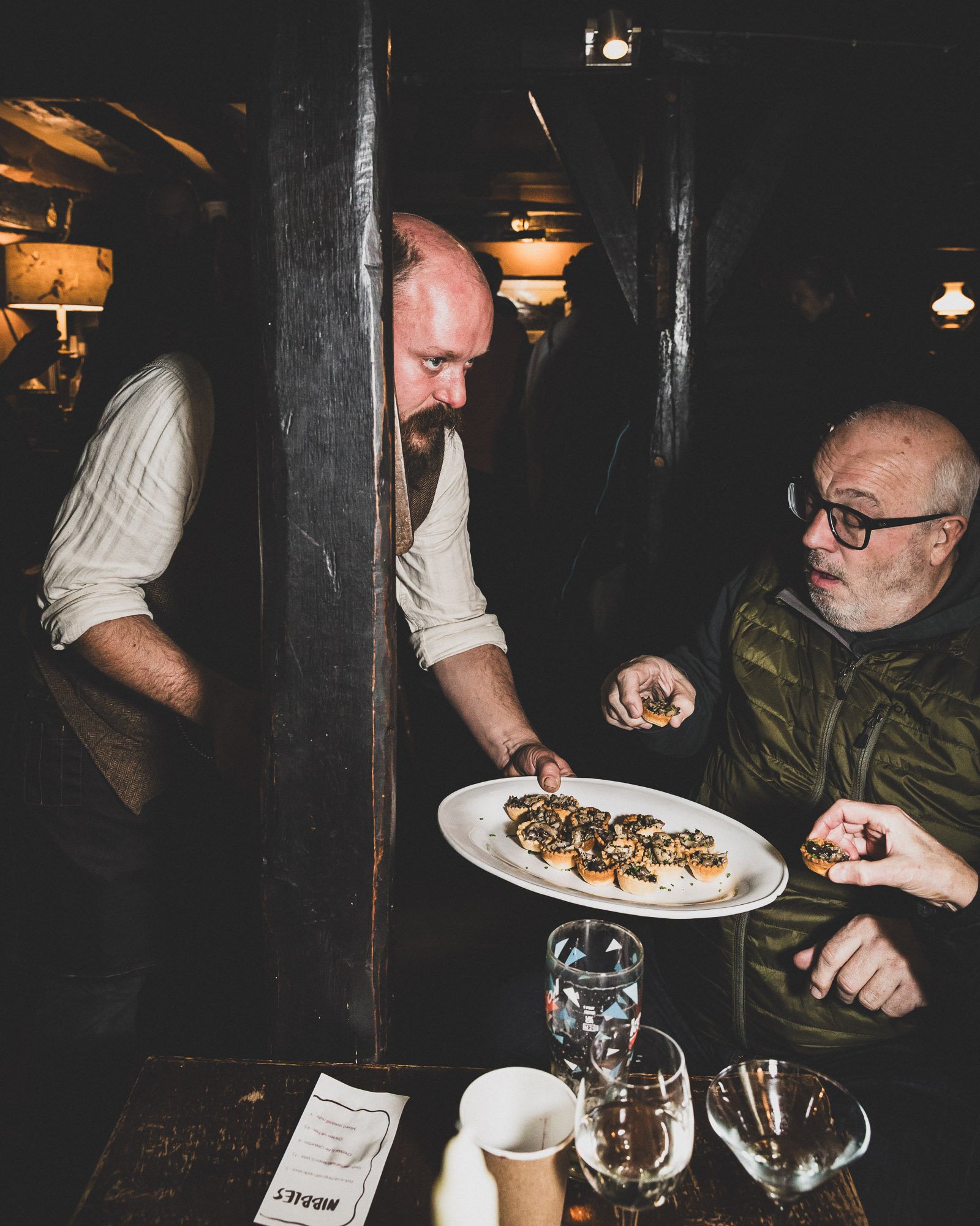 A server, standing behind a wooden partition, is serving a plate of appetizers or canapés to a customer in a dimly lit restaurant or bar. The customer is reaching out to pick up a canapé.