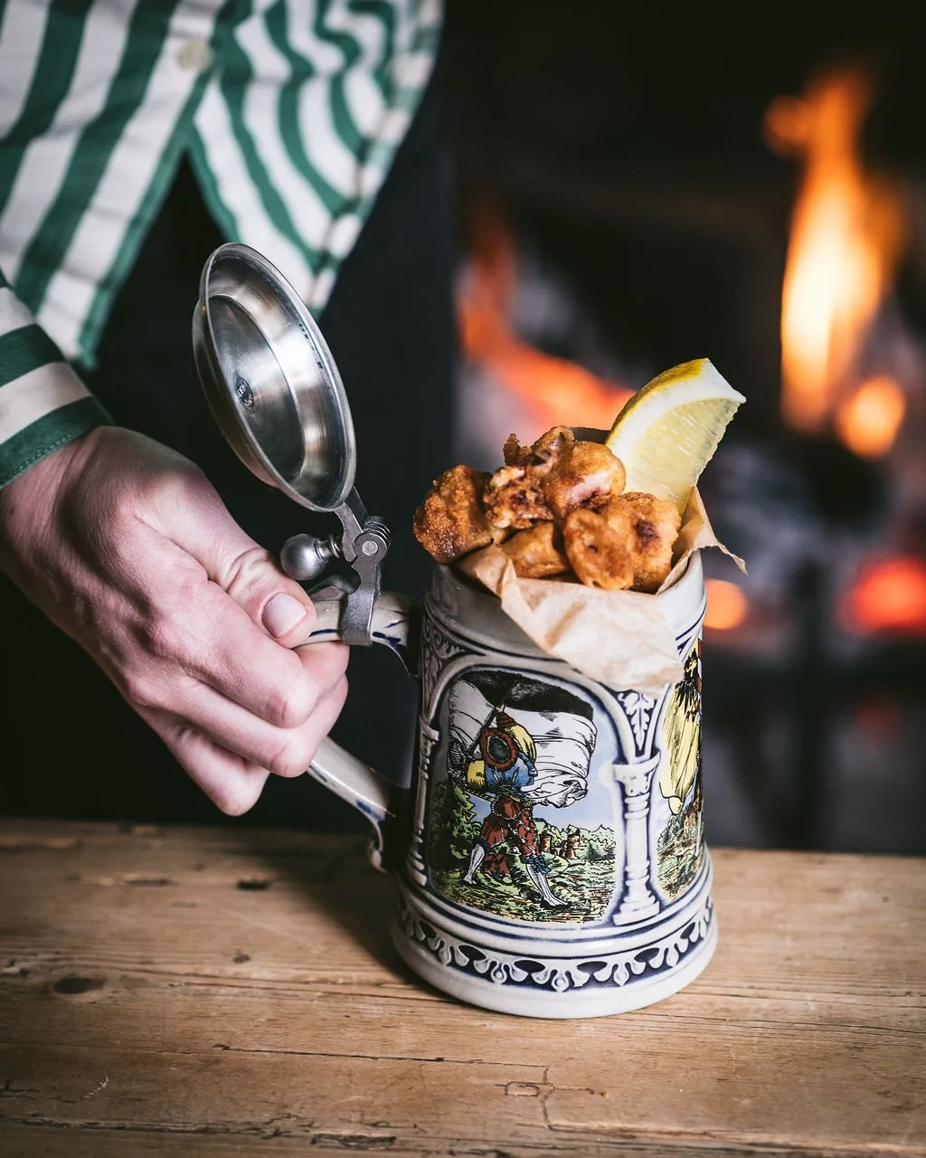 Person holding a metal lid over a ceramic mug filled with fried chicken pieces and a lemon wedge, with a fireplace in the background.