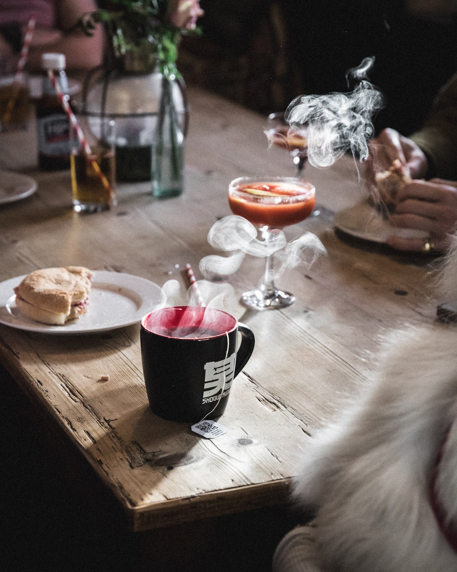 A wooden table with a steaming black coffee mug, a plate with a sliced sandwich, and two drinks with garnishes; one with a straw and the other in a cocktail glass, with a background of miscellaneous items and a person's hand holding a rose.