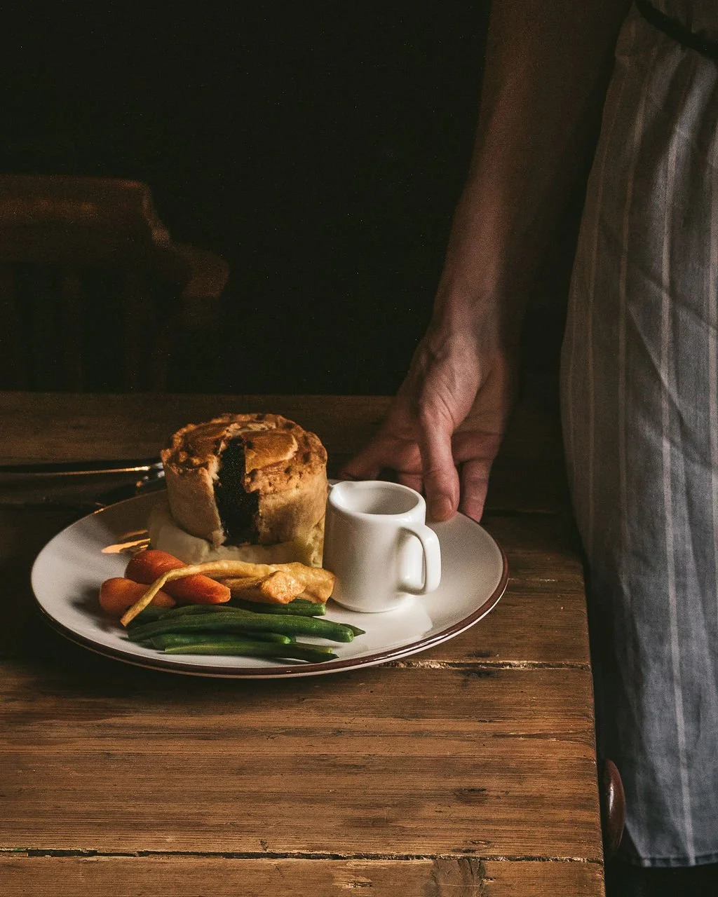 A plate with a partially sliced meat pie, green beans, carrots, and chicken, with a small white jug, on a wooden table. A person in a striped apron is holding the edge of the plate.