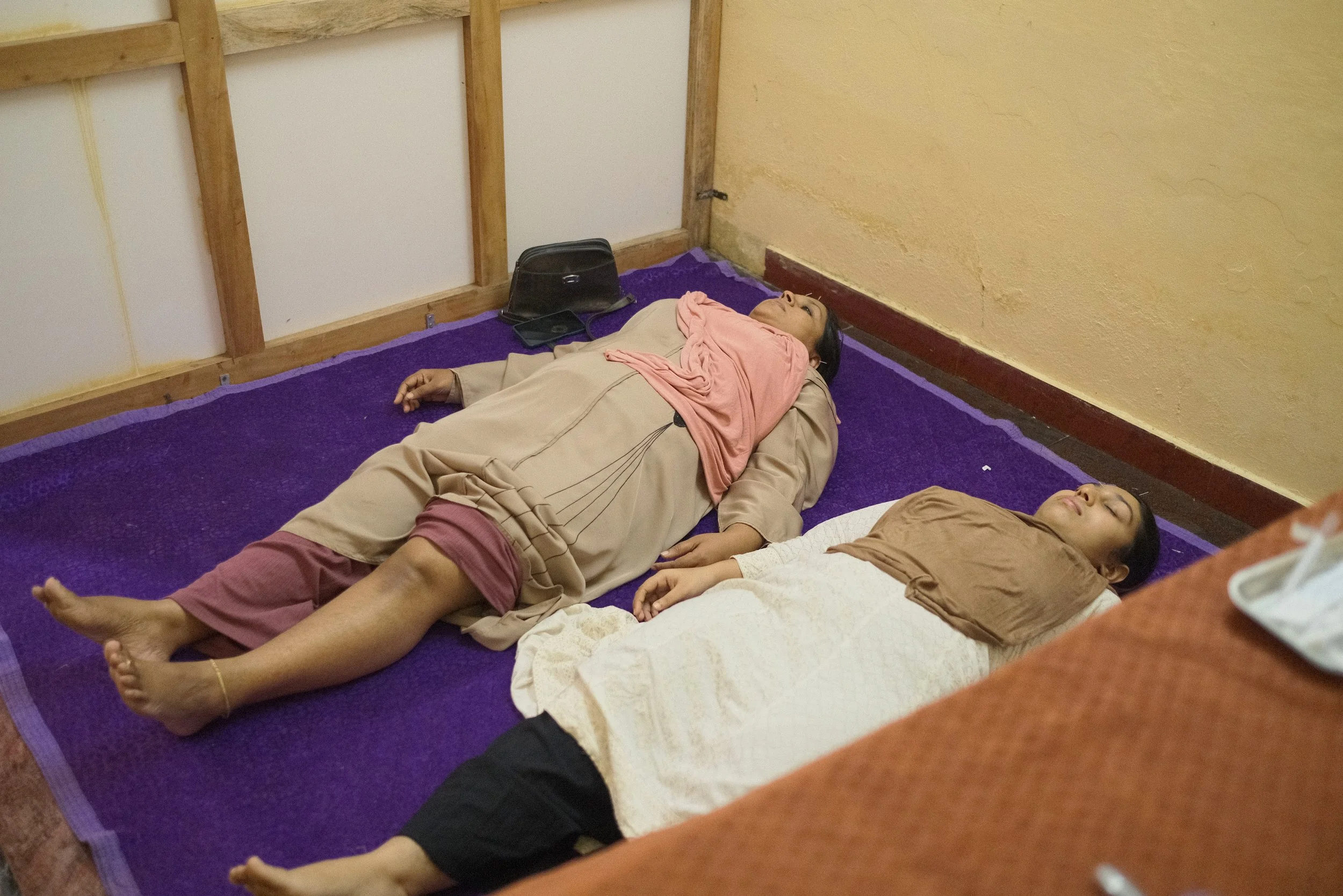 Three women lying on a purple mat with their eyes closed, resting or sleeping in a room with yellow walls and a wooden framed wall in the background.