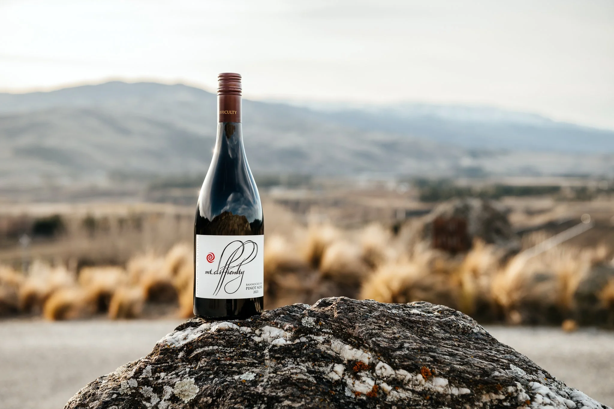 A bottle of Mt. Difficulty wine placed on a large textured rock with a blurred natural landscape background, including mountains and dry grasses.