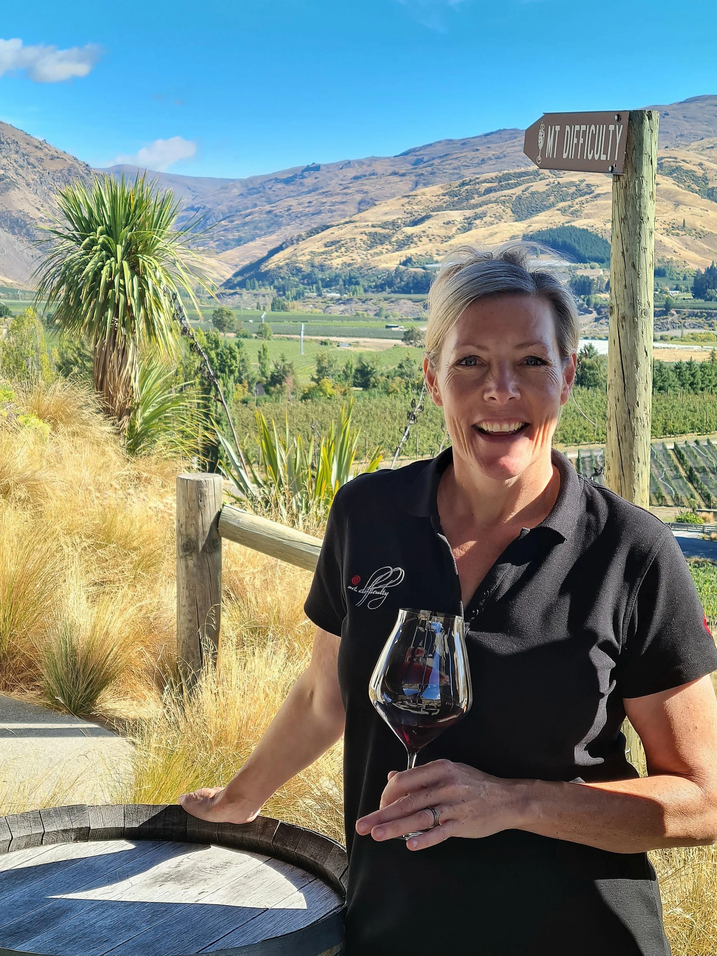 A woman smiling and holding a glass of red wine outdoors with vineyard and mountain landscape in the background.