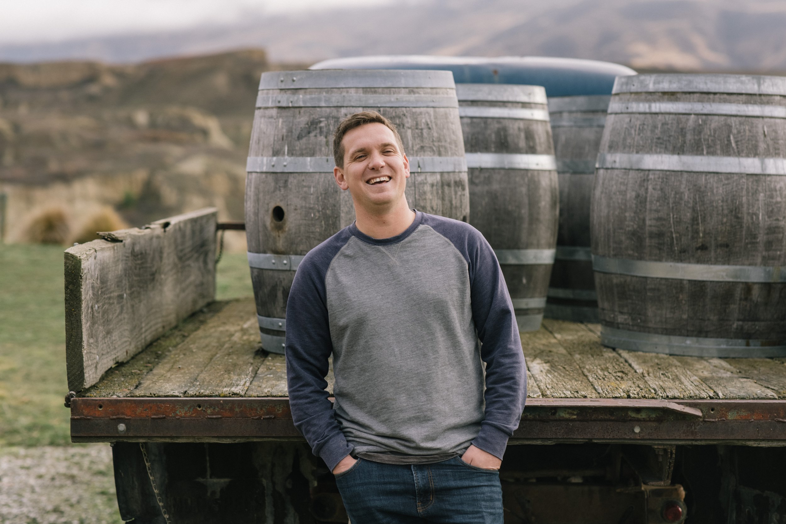 A smiling man standing with hands in pockets in front of stacked wooden barrels on a flatbed truck outdoors.