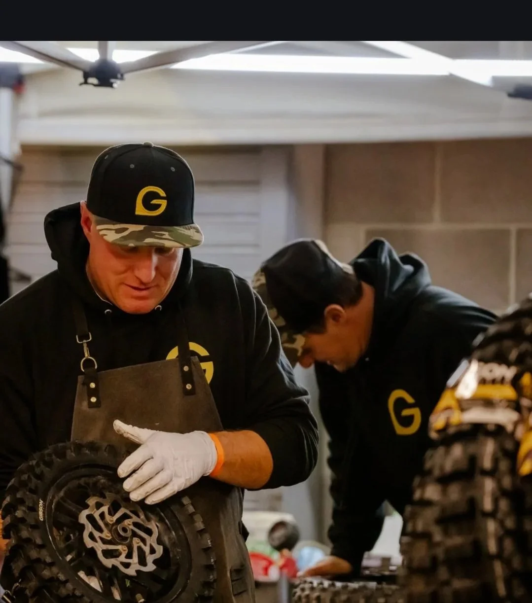 Two men working on a dirt bike engine. One is wearing a black hoodie, a black cap with a yellow G logo, and a gray apron, and the other is wearing a black hoodie with a yellow G logo and a camouflage cap.